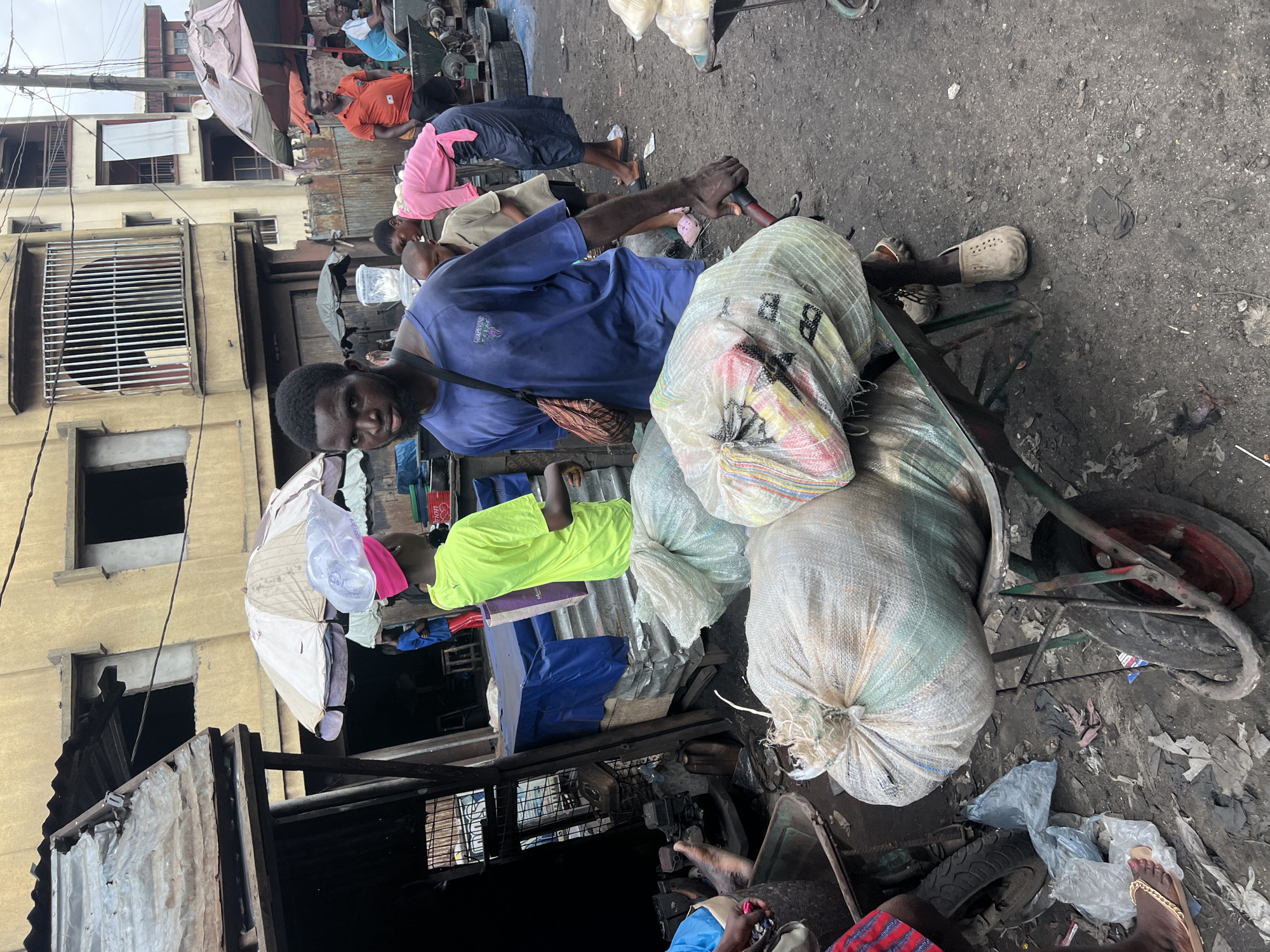 The image captures a vibrant street scene in Nkpor, Nigeria, on an overcast day. The central figure is a young man, appearing to be in his 20s or 30s, with dark skin and a beard. He wears a blue t-shirt, which has faint, largely illegible text on it, possibly including "CAMPUS," and dark pants. He looks directly at the camera with a slight smile while holding the handle of a sturdy handcart.
The handcart is laden with several large, loosely packed white mesh sacks, likely filled with various goods. The letter "B" and other indistinct characters are visible on one of the sacks. The cart has a red wheel. The ground is unpaved, appearing dusty and grey, scattered with plastic bags, discarded items, and general debris, indicating a busy thoroughfare.
In the background, other individuals are visible, contributing to the bustling atmosphere. To the left of the main subject, a person is seen wearing a bright neon green shirt. Further back, another person in an orange shirt stands. Other figures wear pink, light brown, and various colors, suggesting a diverse crowd. A child's foot with pink Crocs is visible near the cart, and a person's bare foot with a gold sandal is partially visible in the bottom right corner.
The backdrop consists of multi-story buildings with light-colored, possibly yellowish, facades. Some windows are open and dark, while others are secured with metal grilles. Makeshift structures, including those made of corrugated metal sheets, are also present, lending an informal, market-like quality to the setting. An open, beige-striped umbrella is visible near one of the buildings, likely providing shade. The overall scene depicts a busy, working-class urban environment.