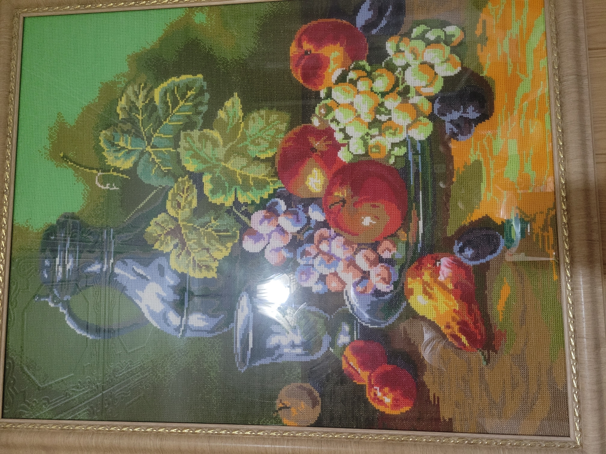 This is a close-up, eye-level photograph of a framed cross-stitch artwork depicting a still life. The artwork is illuminated by a strong light source, causing a significant glare in the center of the image. The subject matter of the cross-stitch is a collection of fruits, including grapes, apples, and what appears to be a pear, arranged around a metallic jug and glass. The background of the artwork is a gradient of green and darker tones. The frame of the artwork is a light-colored wood with a decorative, ornate border. The artwork is hanging on a wall, and the lower portion of the photograph shows a wooden surface, likely a table or floor, with a wood grain pattern. The location of this artwork is not discernible from the image itself; the mention of Belaazyorsk, Belarus, is external information not derived from the visual content.
