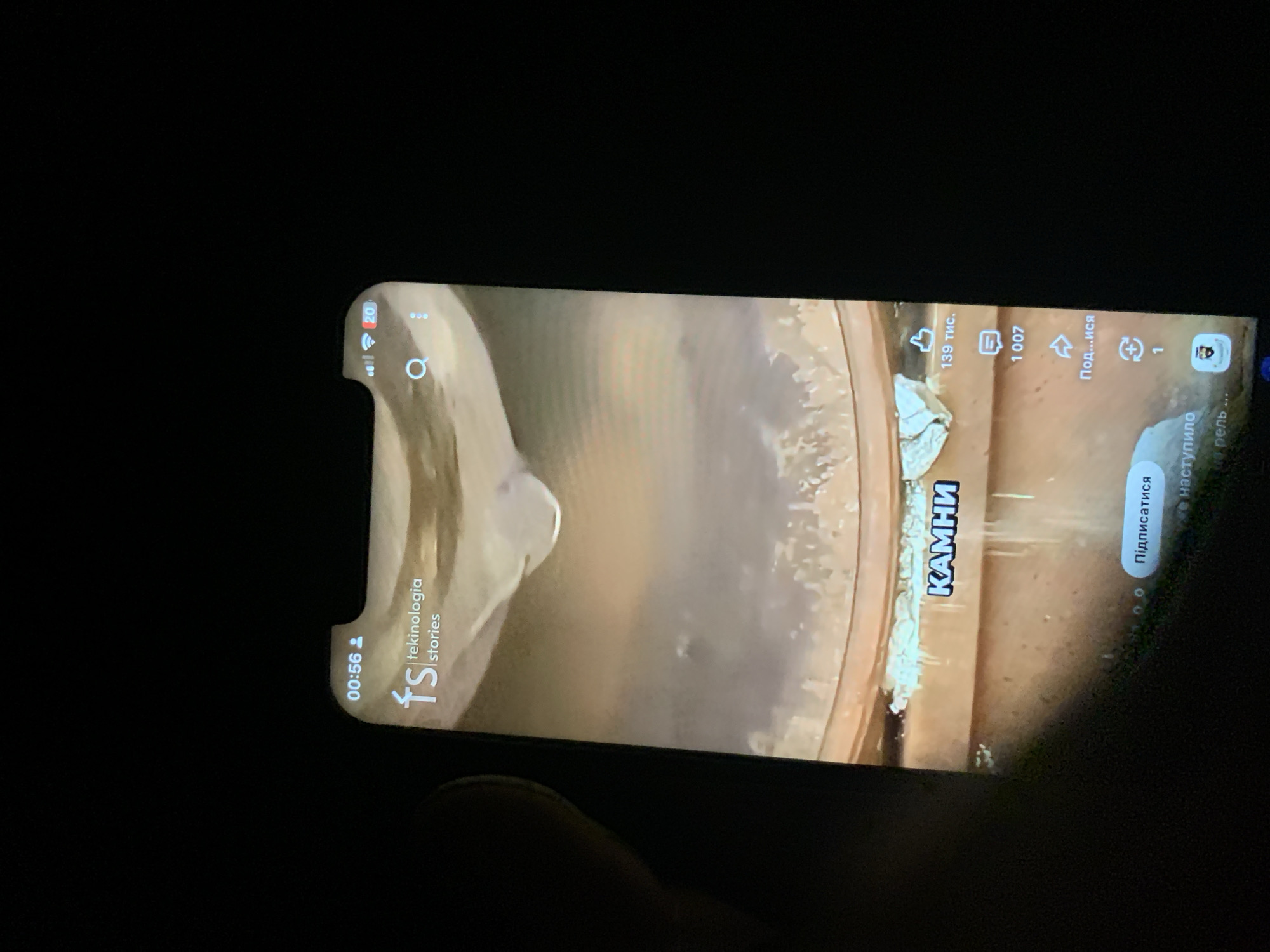 The image displays a smartphone screen in a dark environment, likely taken at night. The phone is displaying a social media video feed, possibly from a platform like TikTok or Instagram Reels. The video itself appears to show a close-up of a substance dripping, with text overlays in what appears to be Cyrillic script.
Key elements visible on the screen include:
* **Time/Status Bar:** At the top left, "00:56" is displayed, likely indicating the video's progress. To the right, icons for signal strength, Wi-Fi, and a notification number (20) are visible.
* **Branding/User Info:** A logo with "fs" and "tekinologia stories" is present on the left side of the screen, suggesting the source of the content.
* **Video Content:** The central part of the screen shows a close-up of a light-colored liquid or substance with a droplet forming. The background appears to be a textured, brownish surface.
* **Text Overlays:**
* "КАМНИ" (likely "stones" in Russian or Ukrainian) is prominently displayed in blue lettering.
* Engagement metrics are shown: "139 тыс." (likely 139 thousand) with a "like" icon, and "1 007" with a comment icon.
* An arrow icon with "Под...ися" (likely "Subscribe" or "Follow") is visible.
* A refresh icon with "1" is below that.
* "Подписаться" (likely "Subscribe") and "наступило" (likely "has come") are also visible.
* **User Interaction Elements:** A subscribe button and potentially other interaction icons are present at the bottom of the screen.
The image is slightly blurred due to the low light conditions and the nature of capturing a screen. The overall impression is that of a user watching a video on their smartphone in a dimly lit setting. There is no identifiable information within the image to suggest a specific location in Zhmerynka, Ukraine, or any other geographical area.