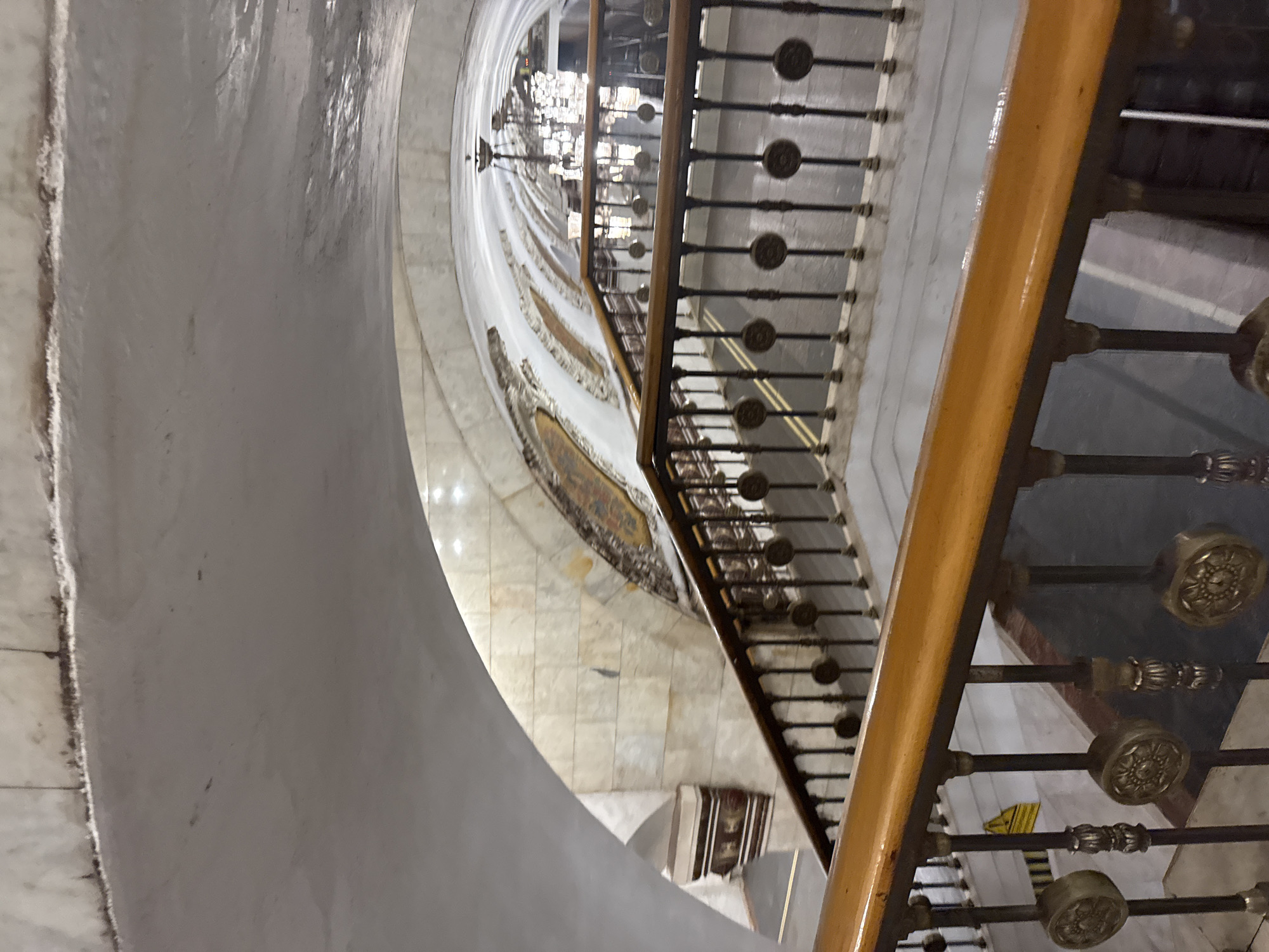 This is an interior shot of a metro station in Moscow, Russia. The image shows a descending staircase with a ornate metal railing and wooden handrail. The walls of the station are clad in marble, and the arched ceiling features decorative elements. Several chandeliers are visible in the background, illuminating the vaulted space. The scene appears to be deserted.