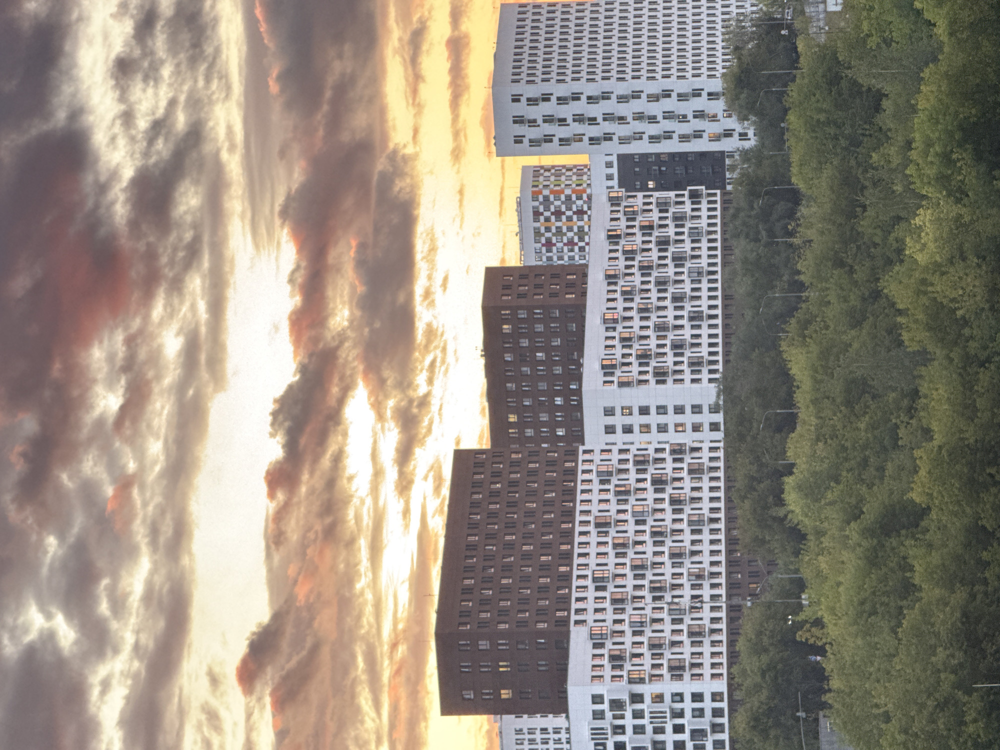 The image shows a collection of modern, multi-story buildings against a dramatic sunset sky filled with colorful clouds. The buildings are a mix of white and dark brown facades, with many windows visible. One building features a colorful, abstract geometric pattern. In the foreground, a dense canopy of green trees obscures the lower portions of the buildings. The scene appears to be an urban residential area. This view is characteristic of the Moscow City area in Moscow, Russia, known for its contemporary architecture and high-rise developments. There are no people or specific actions depicted in the image.