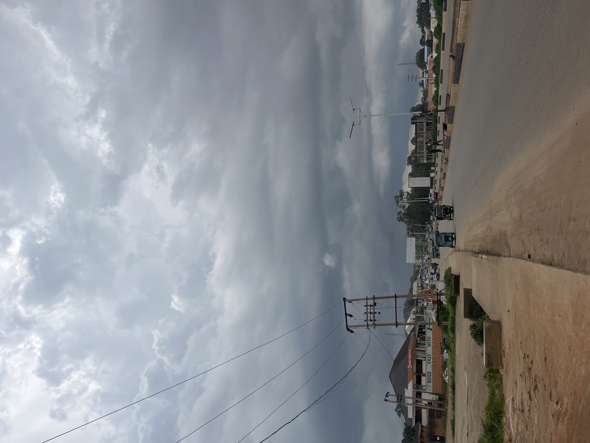 The image depicts a road scene in Gura Top, Nigeria, under a cloudy sky. The sky is filled with heavy, gray clouds, suggesting an overcast or potentially rainy day. The road is paved and appears to be a main thoroughfare, with a concrete sidewalk or median running alongside it.
Several vehicles are visible on the road, including cars, motorcycles, and a three-wheeled auto-rickshaw (likely a Keke Napep). Some people are seen walking along the sidewalk and near the road. Buildings line the side of the road, some appearing to be under construction with exposed brickwork and scaffolding. One building prominently displays text on its side: "BUILD EASY WITH." Other smaller buildings and some trees are visible in the background, with more structures and a power transmission tower further in the distance.
The overall impression is of a typical day in a Nigerian urban or semi-urban setting, characterized by traffic, construction, and the presence of everyday infrastructure. The cloudy weather lends a somewhat subdued atmosphere to the scene. The image is oriented vertically, with the road and buildings running from bottom to top on the right side of the frame, and the sky dominating the left.