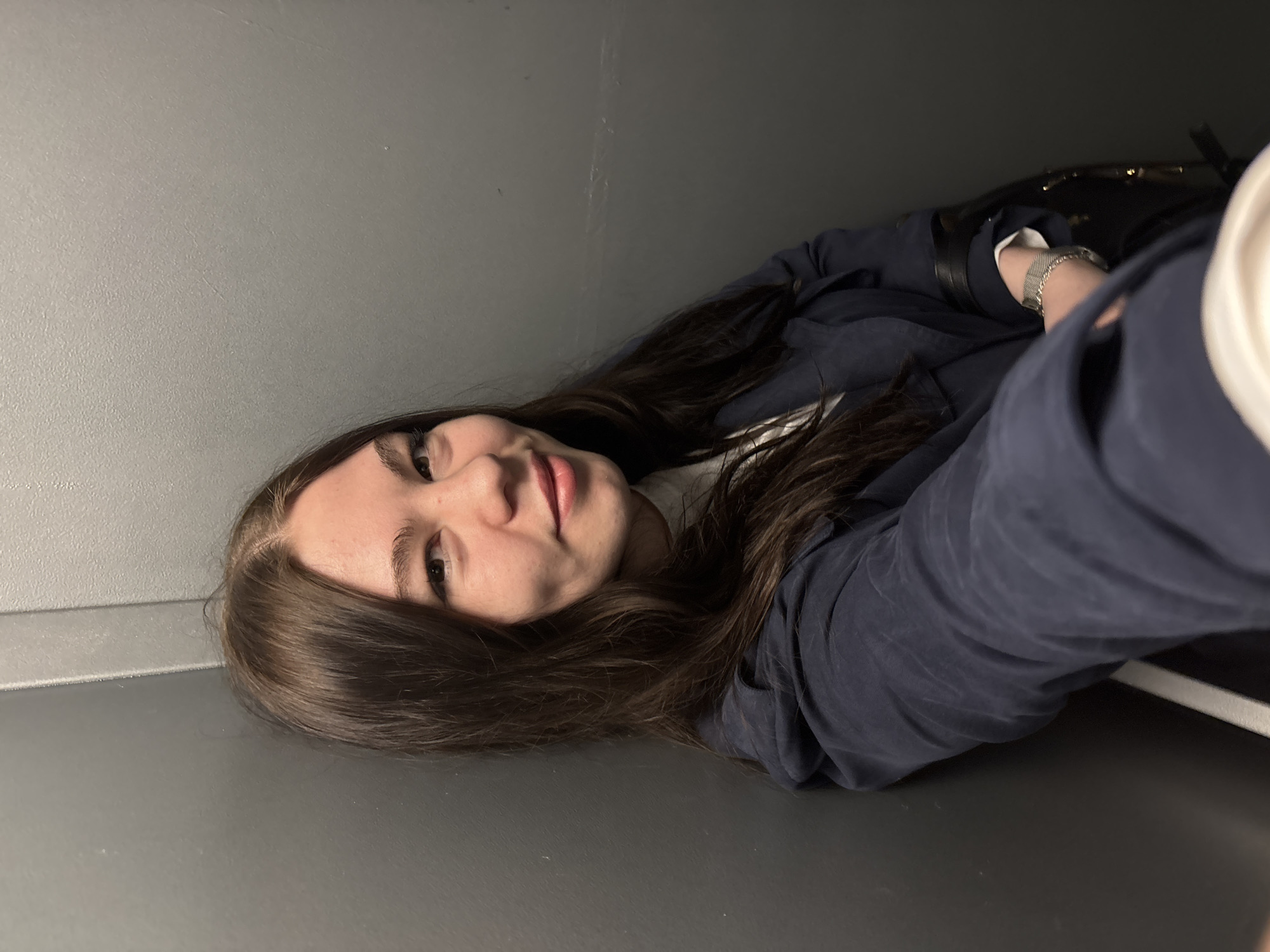 A young woman with long brown hair is lying on a gray surface, possibly a floor, with a gray wall behind her. She is wearing a dark blue jacket and a silver watch. A dark bag is visible in the background. The location is not discernible from the provided image.