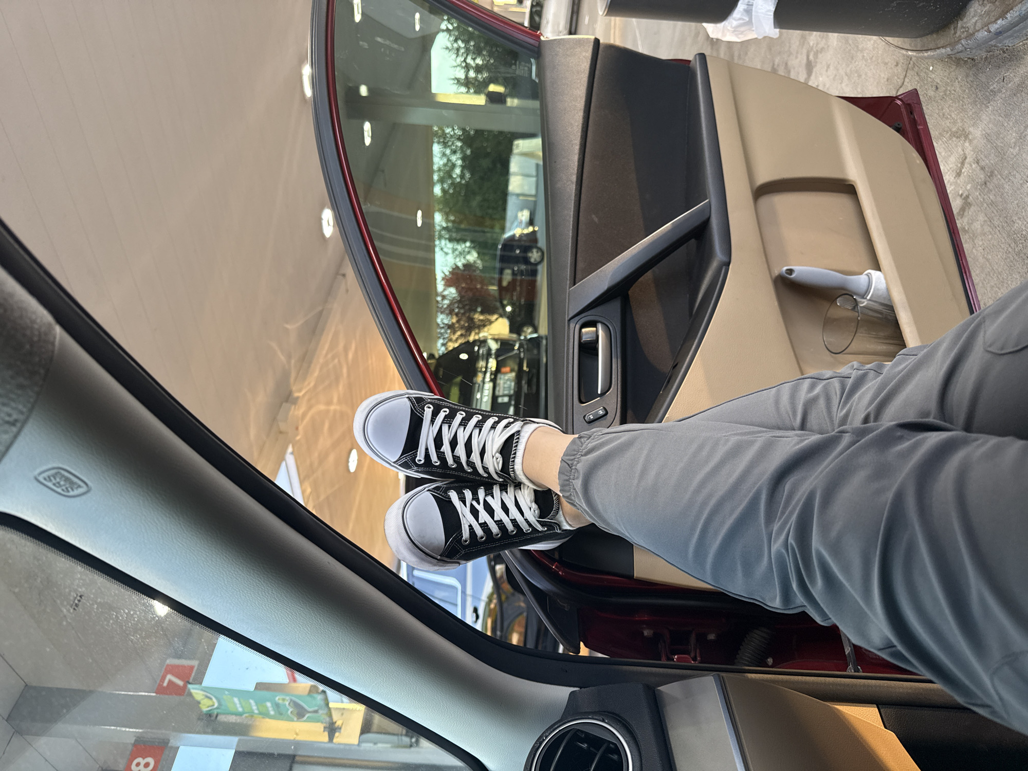 The image shows a person's legs and feet, wearing black high-top sneakers with white laces, propped up on the open car door of a dark red vehicle. The person is wearing grey sweatpants. The scene appears to be at a gas station in Fife, United States of America, with gas pumps and signage visible in the background through the car windows. The interior of the car shows the door panel with a handle and a storage compartment containing a glass and a lint roller. The lighting suggests it is either early morning or late evening.