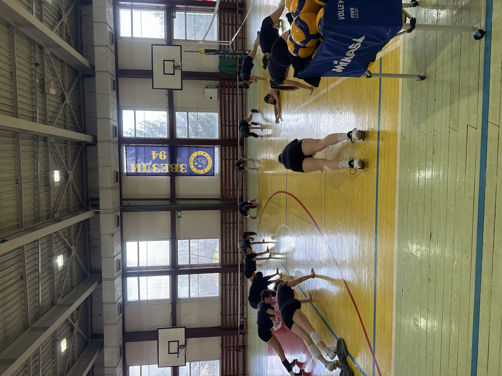 A gymnasium in Sofia, Bulgaria contains a volleyball team stretching on a court. The players, all wearing black tops and shorts, are in a line performing a dynamic stretch. Windows line one side of the gym near a banner which says "3 ЗВЕЗДИ 94." Volleyballs are in a cart near a volleyball net. The court lines are visible on the wooden floor, which has a light-yellow and light-green color. Basketball hoops hang on the wall.