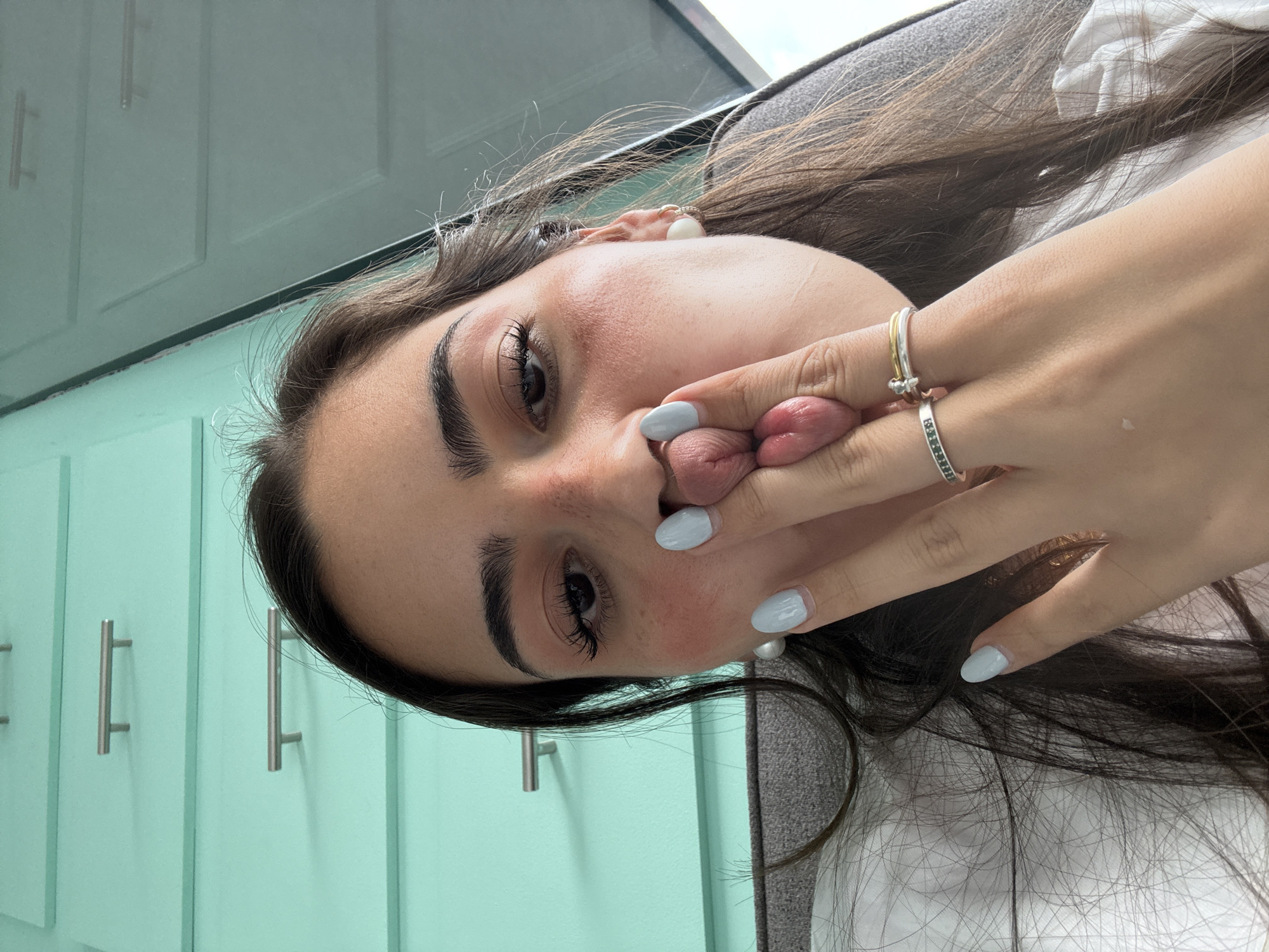 A woman with long brown hair, wearing rings on her hand, is holding her tongue with her fingers, posing for a selfie. She has fair skin, dark eyebrows, and wears earrings. She is likely indoors, with light blue cabinets visible in the background. The specific location, San Bernardino Tlaxcalancingo, Mexico, cannot be determined from the image.