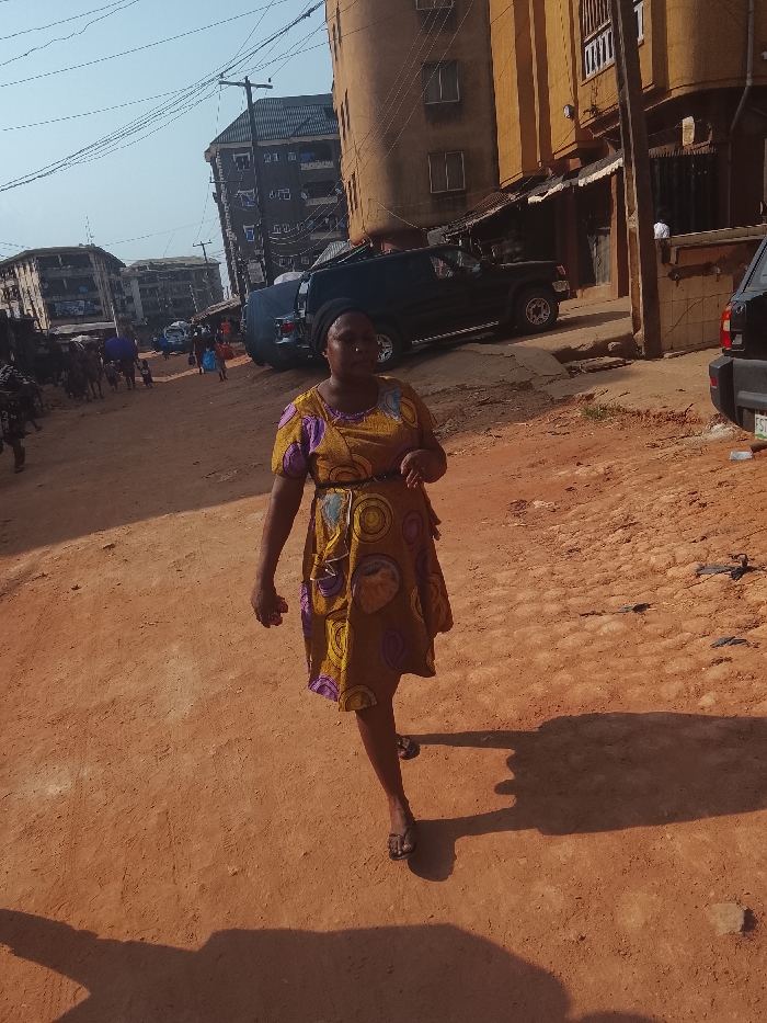 A woman in a patterned dress walks along a dirt road in an urban setting. Buildings line the street, and vehicles are parked nearby. The road appears uneven and somewhat worn. The woman casts a long shadow on the ground, suggesting a low angle of sunlight. Other people are visible in the background, adding to the scene's busy atmosphere.