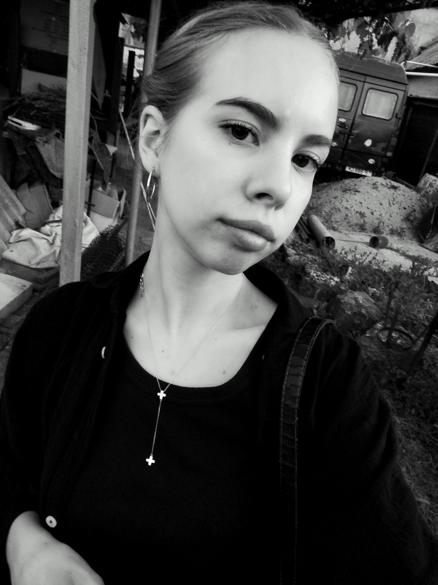 In a black and white, medium shot, a young woman with her hair pulled back in a bun is seen from the chest up, taking a selfie outdoors. She is wearing a dark, possibly black, long-sleeved shirt, a necklace with two cross pendants, and hoop earrings. She is standing in front of a pile of construction debris, including lumber and possibly sheeting, to her left. Behind her and to her right is a mound of dirt with some vegetation and a dark-colored van. The overall environment appears to be a somewhat disheveled outdoor area, possibly a construction site or a yard with stored materials. The lighting is dim, contributing to the grayscale aesthetic.