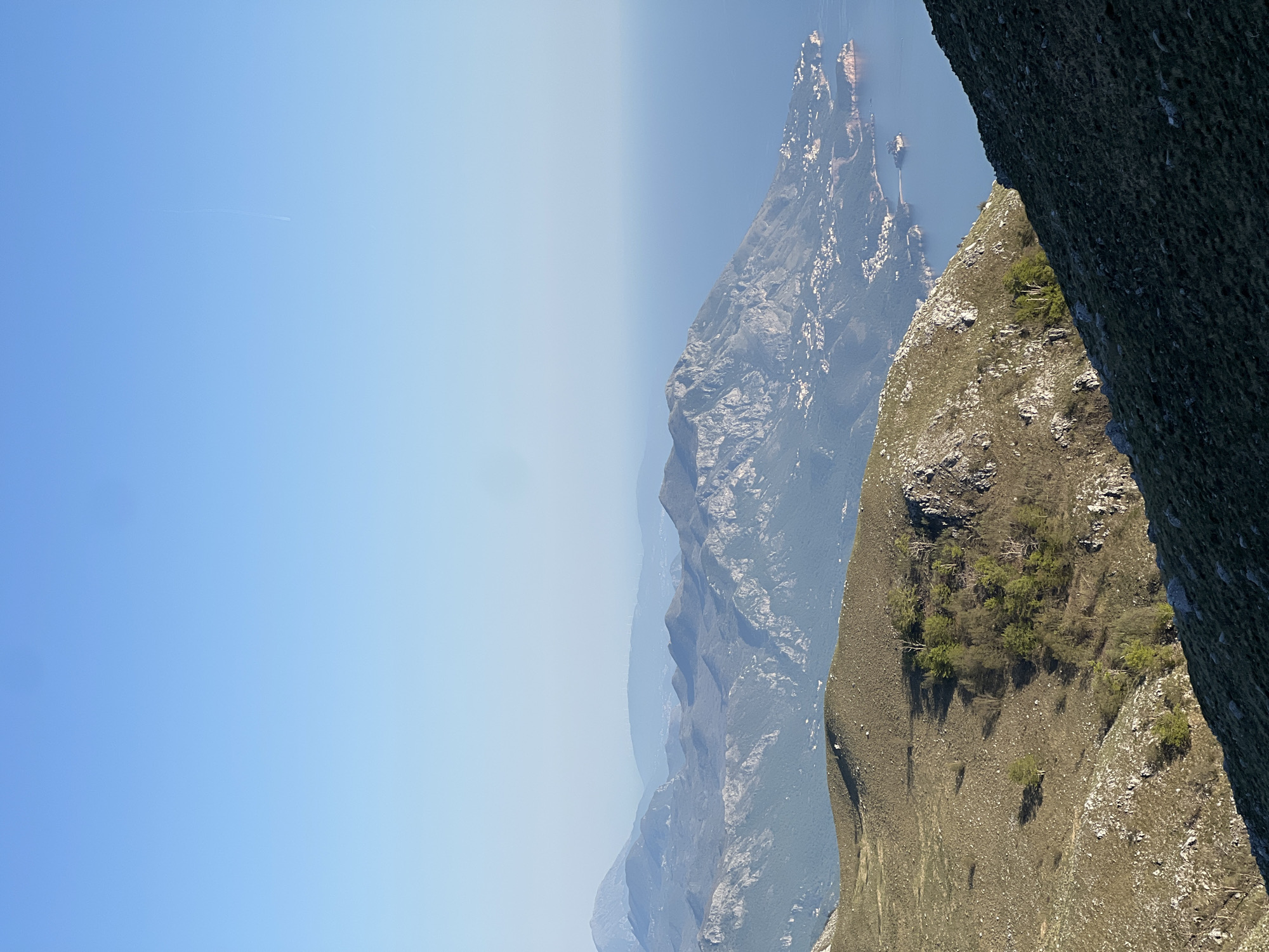 The image shows a view of mountains and a body of water in Dolovi, Montenegro. The sky is clear blue with a contrail visible on the left side of the image. Mountain ranges stretch into the distance, becoming hazy. On the right side of the image, there is a cliff face with grass and some small trees growing on it.