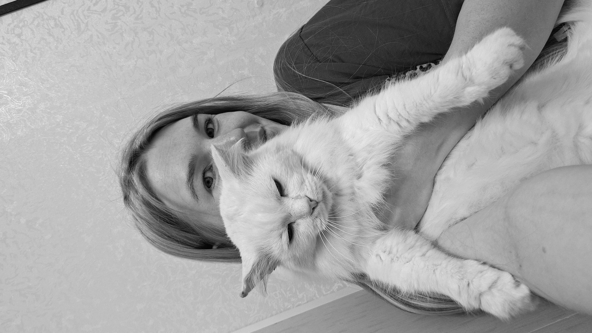 A black and white, eye-level, medium shot shows a woman holding a fluffy white cat. The woman's face is partially obscured by the cat, but her eyes are visible and she is smiling. The cat is looking down with its eyes partially closed. The background is a textured wall. The image does not contain information about the location within the city of Большой Букор, Russia.
