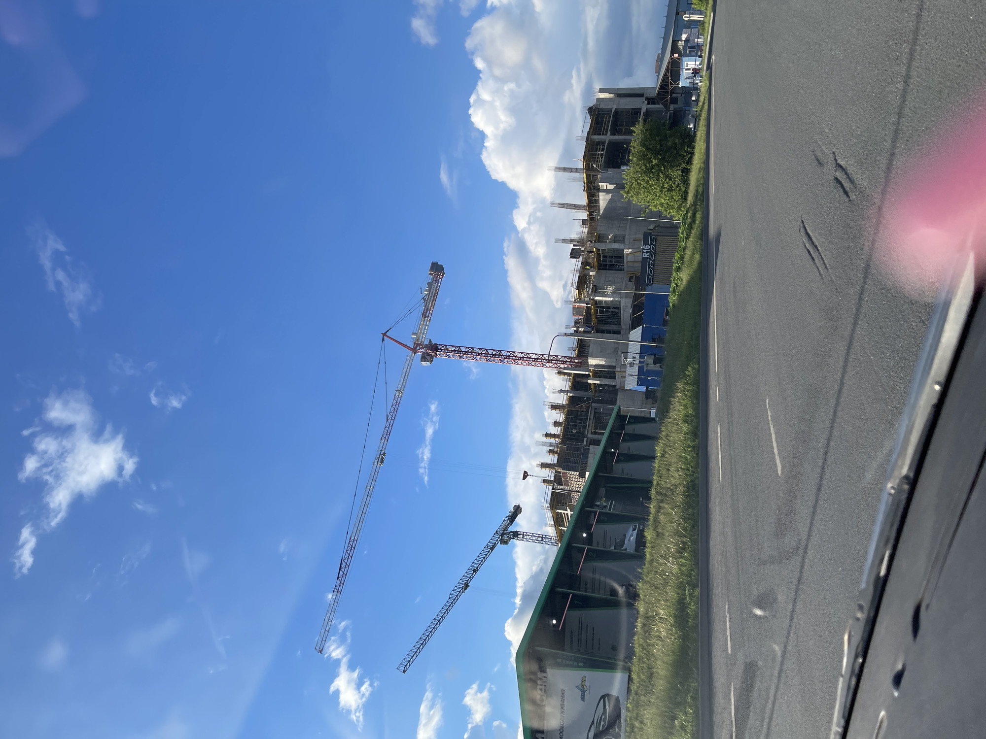 The image shows a construction site in Bittsa, Russia. The scene features unfinished buildings with multiple cranes extending into a bright blue sky with scattered clouds. There's a roadside visible, and some greenery near the construction site. The photograph was taken from inside a vehicle, as part of the car's interior is visible, including a portion of the dashboard and a pink reflection on the right.