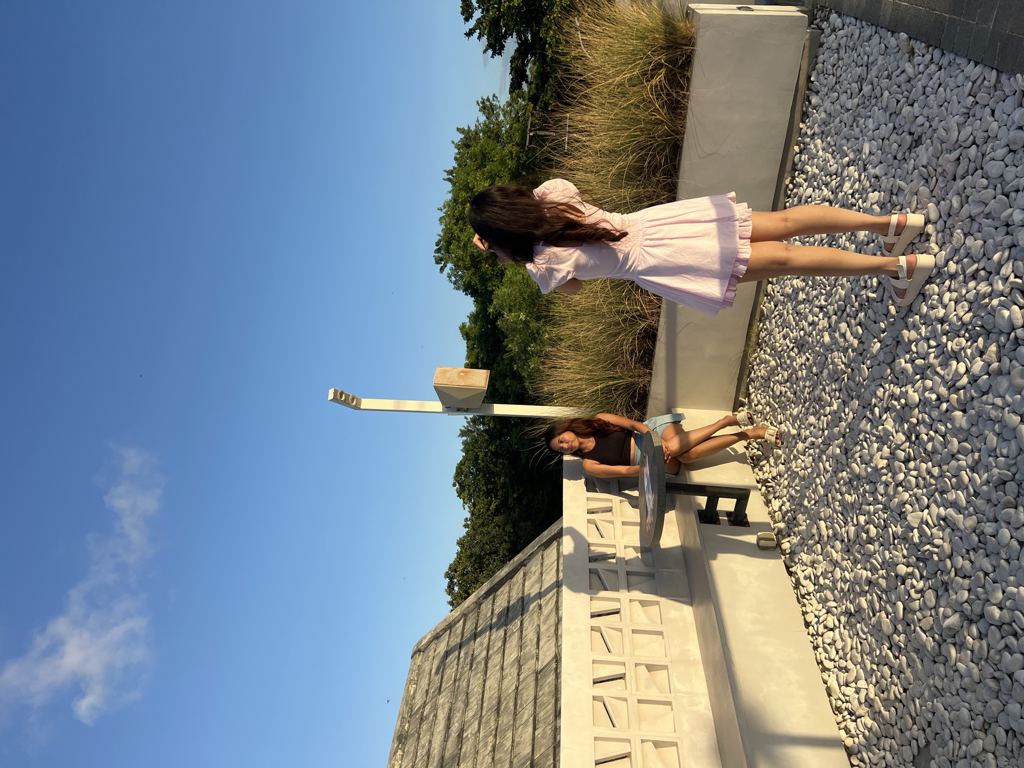 The image depicts two women in an outdoor setting. One woman, wearing a pink dress and light-colored shoes, stands on a surface covered in white stones. She is facing away from the camera and appears to be looking out at the scenery. The other woman is seated on a concrete structure that includes a table or counter. She is wearing a dark top and light-colored shorts. Behind them is a mix of vegetation, including trees and tall grass, set against a clear blue sky. The location might be Kuta, Indonesia due to the appearance of tropical vegetation and architecture.