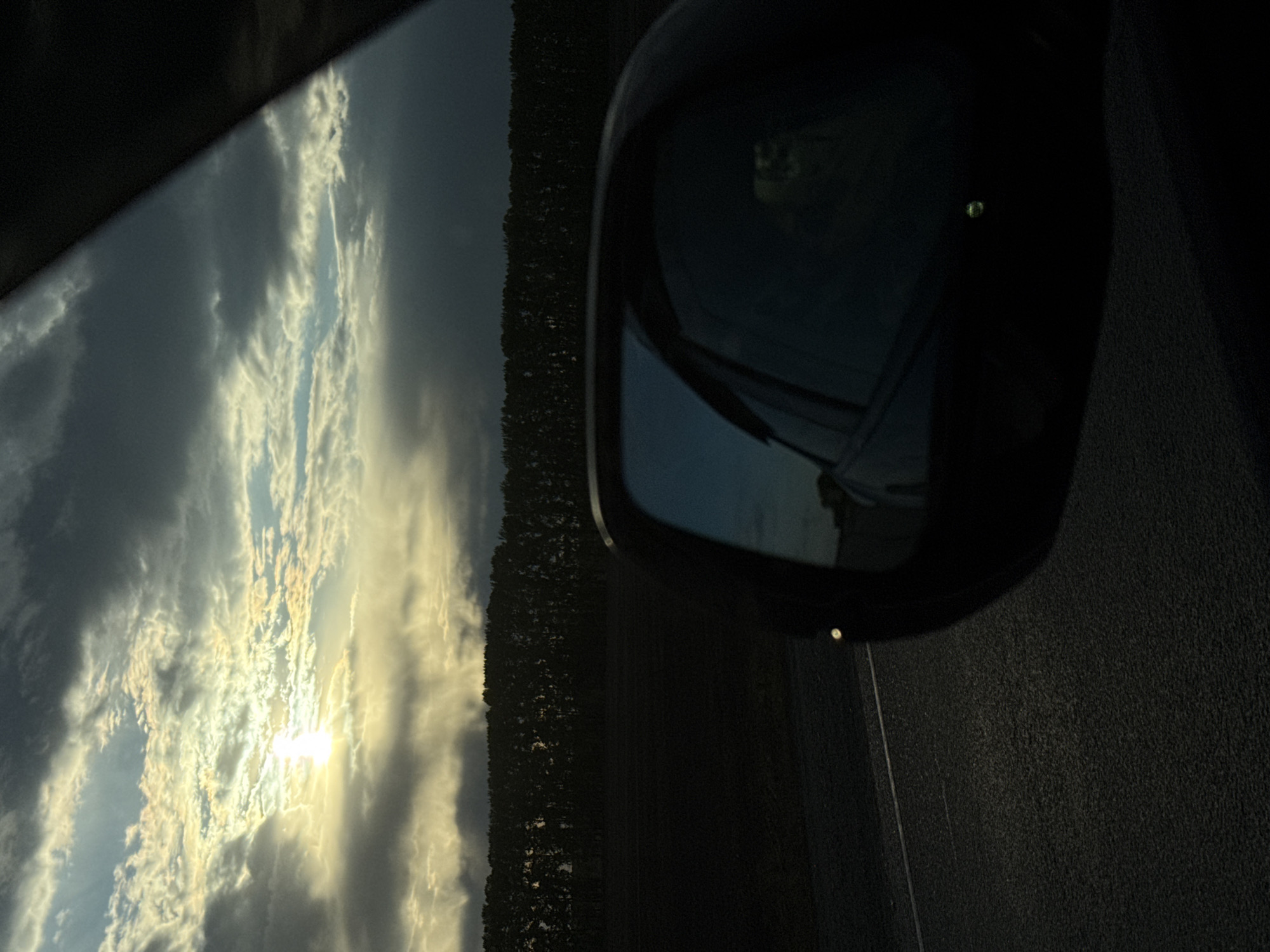 The image depicts a view from a car's side mirror, showing a landscape under a cloudy sky. The sun is partially obscured by clouds, creating a bright glow. A dark strip of trees lines the horizon. The road is visible in the foreground, appearing dark. The location is Unknown, Unknown.
