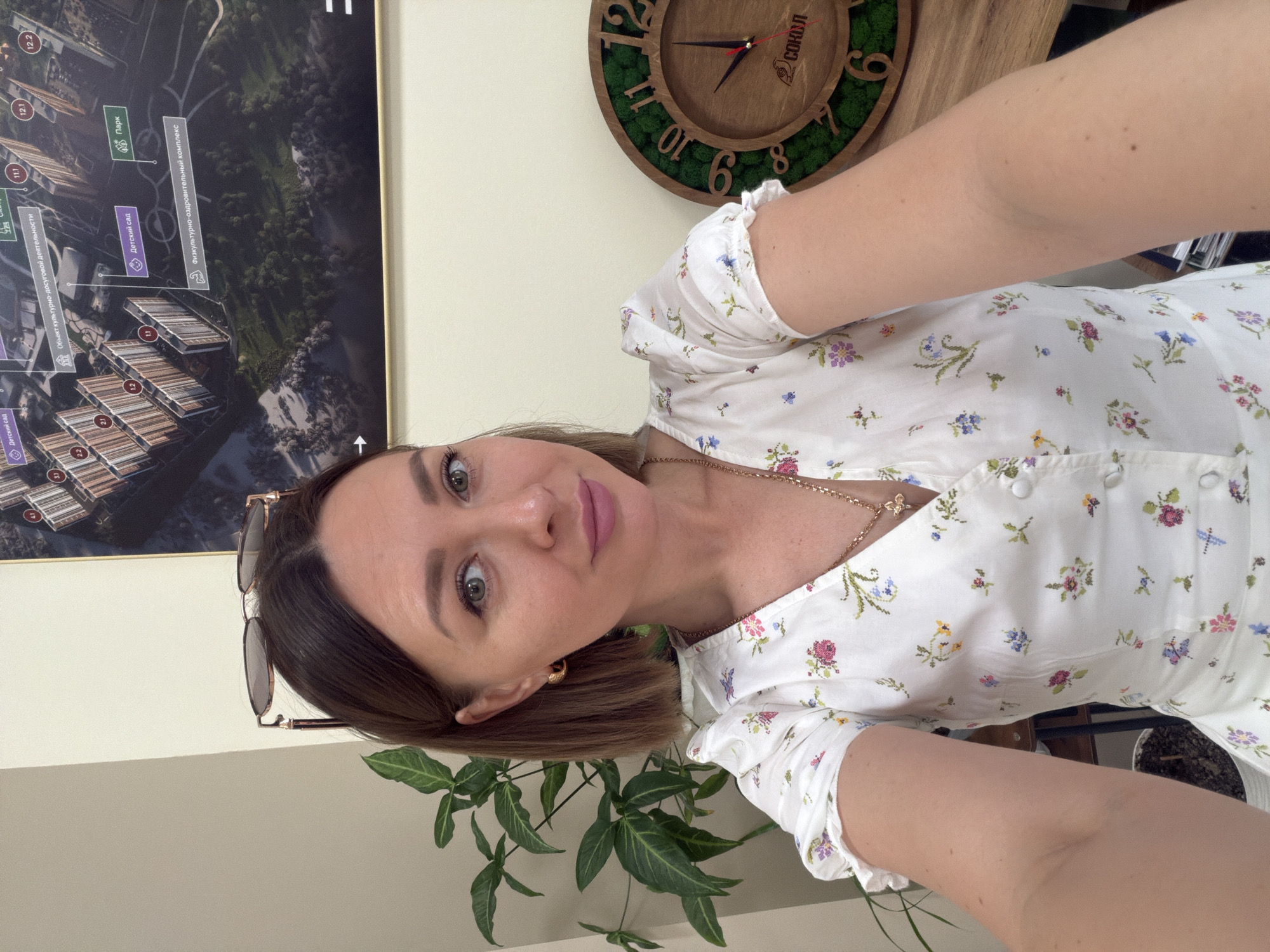 Here is a detailed description of the image:
A woman is posing for a selfie in what appears to be an indoor environment. The camera is angled slightly downward towards her, capturing her upper body and face. She is wearing a white floral-patterned dress with short, ruffled sleeves. A gold necklace is visible around her neck. Her short brown hair is styled neatly, and she is wearing sunglasses propped on her head.
In the background, there is a framed aerial view of a planned neighborhood which includes various buildings and green spaces. Text in the background labels the aerial view in Russian, stating "Children's Garden", "Park" and "Cultural and Leisure Activities" as well as "Sports and Recreation Complex". There is also a round wooden clock with green moss accents on the wall. A portion of a green leafy plant can be seen at the bottom of the frame.
Based on the details in the aerial view picture, it seems to be located in Rostov-on-Don, Russia.