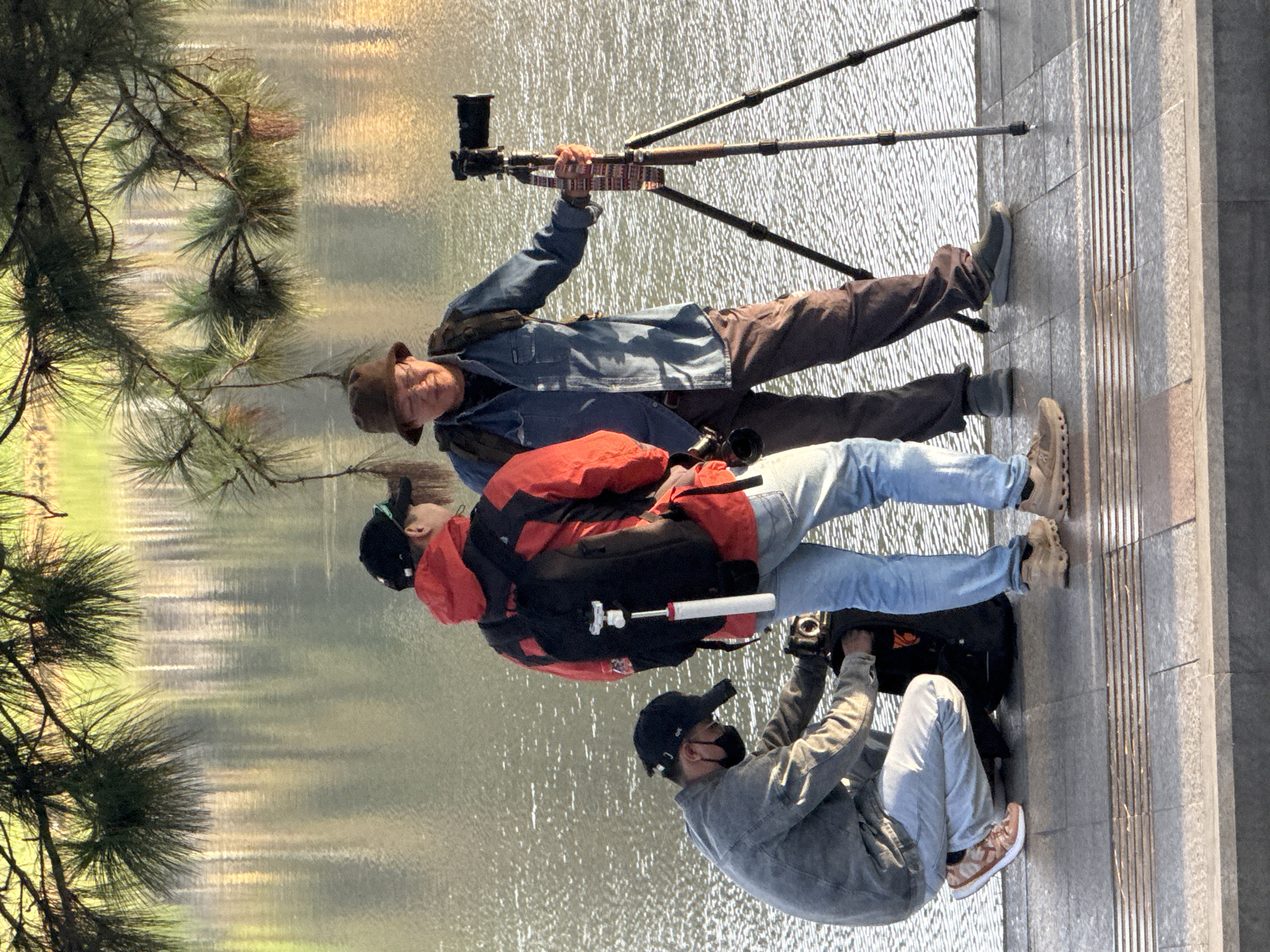 Three individuals are present by a body of water, likely Xuan Huong Lake in Da Lat, Vietnam, given the context. In the foreground, one person wearing a grey jacket, white pants, a black baseball cap and black face mask is crouched down, reaching into a black backpack. To their right, another person in a red and black jacket and blue jeans stands holding a camera. Further to the right, a third person wearing a denim shirt, brown pants, and a brown hat is holding a camera mounted on a tripod. The water behind them reflects the surrounding trees and sky, creating a shimmering effect. The scene appears to be a waterfront promenade, with tiled ground in the foreground and foliage from overhead trees casting shadows. The lighting suggests it is late afternoon or early morning.