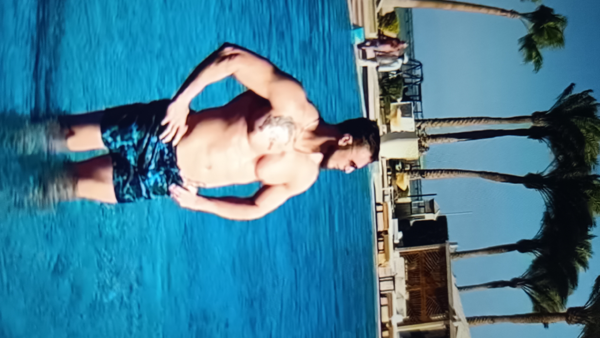 The image shows a person in a swimming pool, with palm trees and a building in the background. The person is a man, shirtless and wearing patterned swim trunks. He is standing in the water, with his arms outstretched. The water is a vibrant blue and appears to be clear. In the background, there are several palm trees with green fronds, and a building with a light-colored exterior. The sky is a clear blue. The scene appears to be a tropical or resort setting.