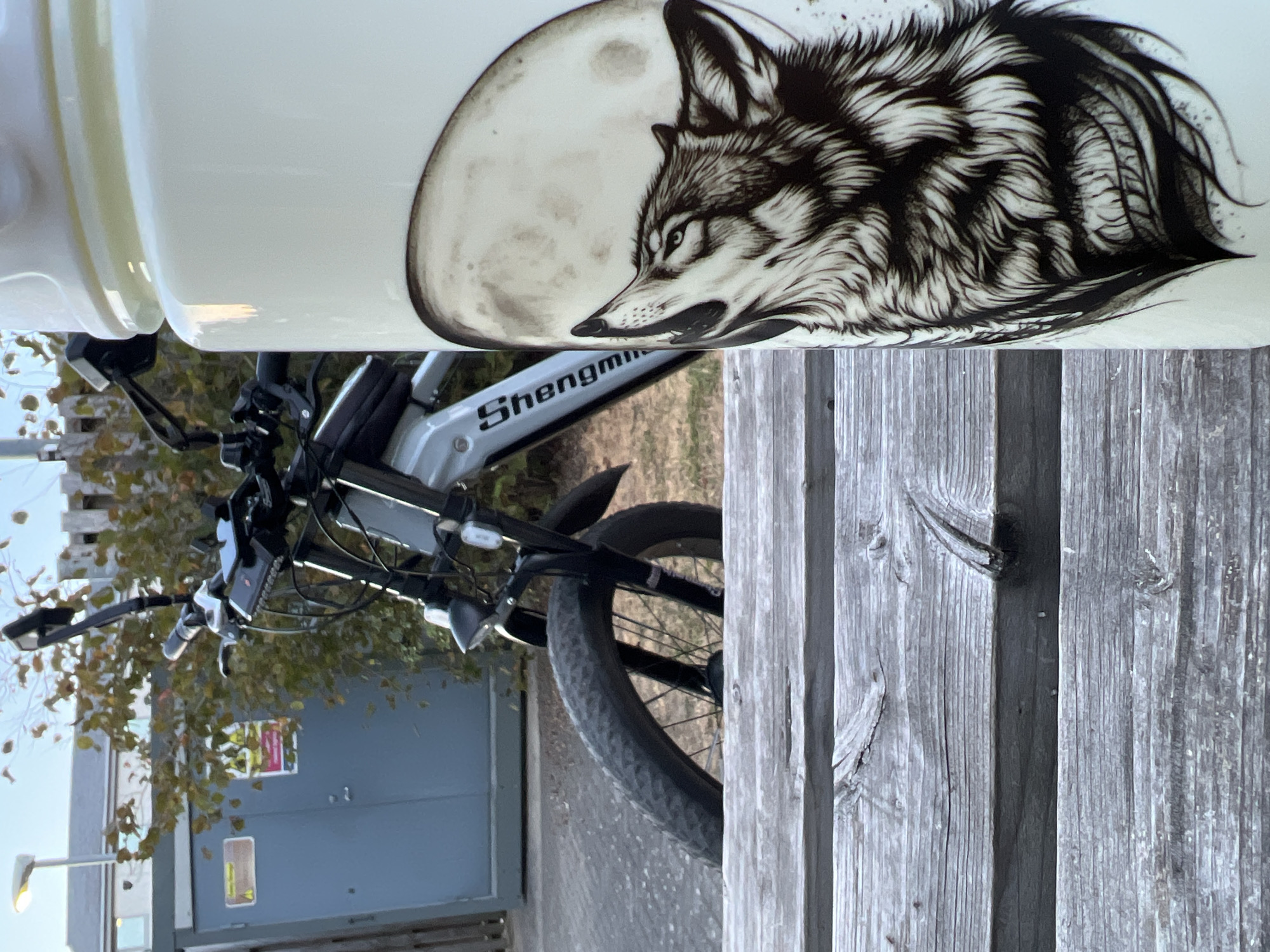 The image is an outdoor close-up shot of a white thermos bottle with a black and white illustration of a wolf howling at a full moon. The thermos is placed on a weathered wooden bench in the foreground. In the background, a silver electric bicycle is partially visible. The bicycle has a black frame, thick tires, and a black bag attached to the handlebars. Behind the bicycle is a grey metal utility box and some green foliage. The scene appears to be set in an urban or suburban environment. The overall lighting suggests it is either early morning or late afternoon. There are no people present in the image. The image does not provide any specific information to determine the location as Guildford, United Kingdom.