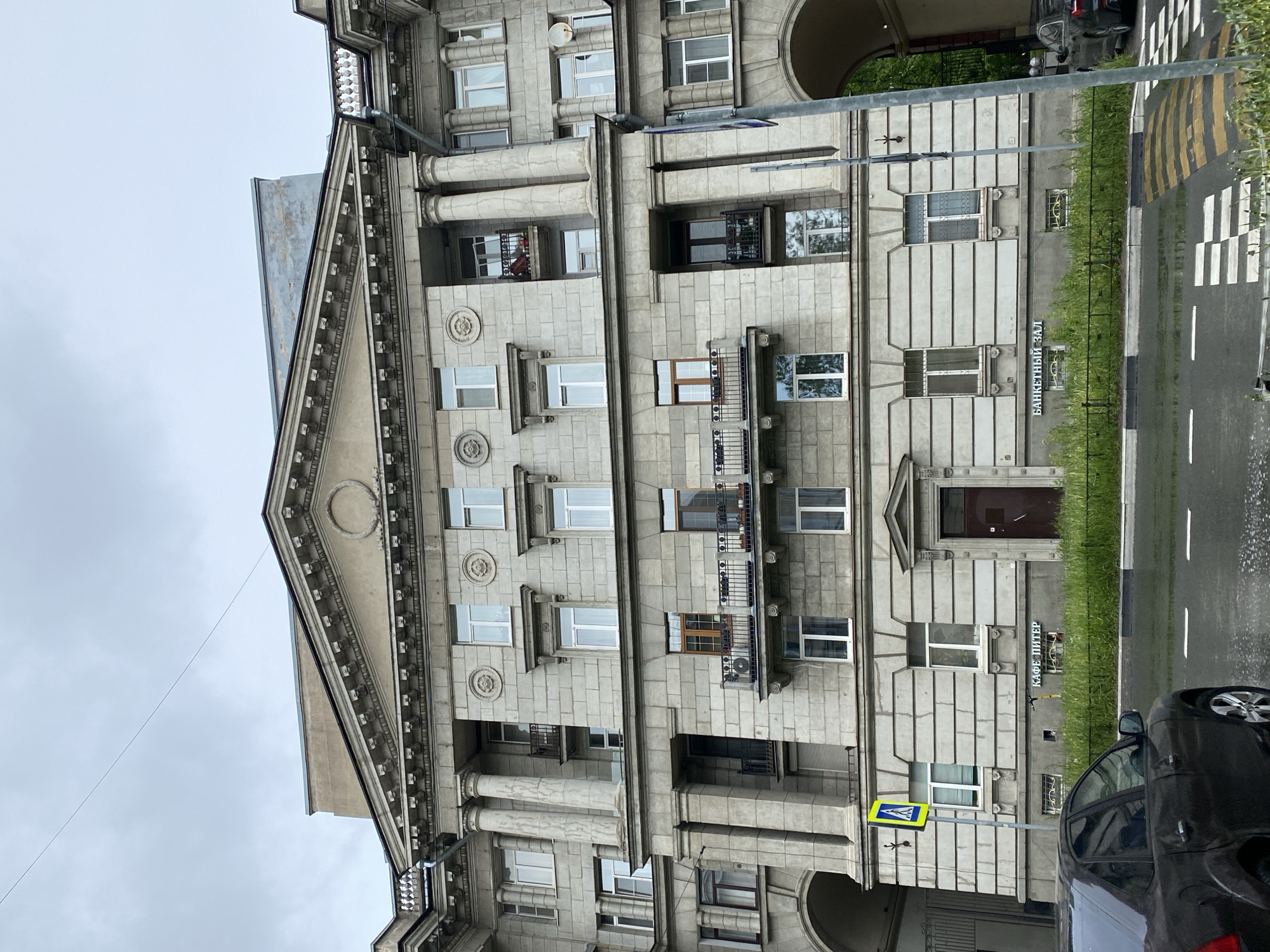 The image shows the facade of a building in Saint Petersburg, Russia. It's a grey, stone-faced building with architectural details like columns, decorative trim, and a triangular pediment at the top. The building has multiple windows and balconies. A sign reading "БАНКЕТНЫЙ ЗАЛ" (Banquet Hall) is visible in front of the building, next to which is another sign "КАФЕ ПИТЕР" (Cafe Peter). A pedestrian crossing sign and a zebra crossing road marking are visible near the building. A portion of a car is in the foreground. The sky is overcast.