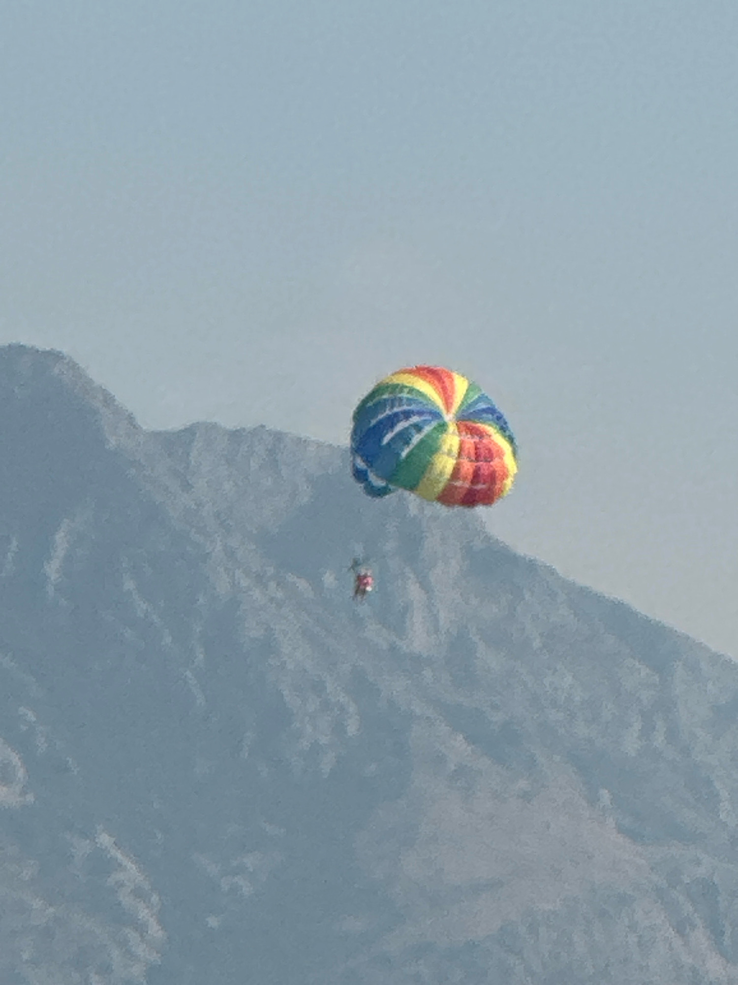 The image shows a rainbow-colored parachute with a person harnessed underneath, parasailing in the sky. The parachute is positioned in the upper right quadrant of the image, while a large, hazy mountain range dominates the lower portion. The sky is a pale blue and appears to be slightly overcast or hazy. The overall scene suggests an outdoor recreational activity taking place in a mountainous coastal area. Without additional information, the specific location in Boreti, Montenegro, cannot be confirmed from the image alone.