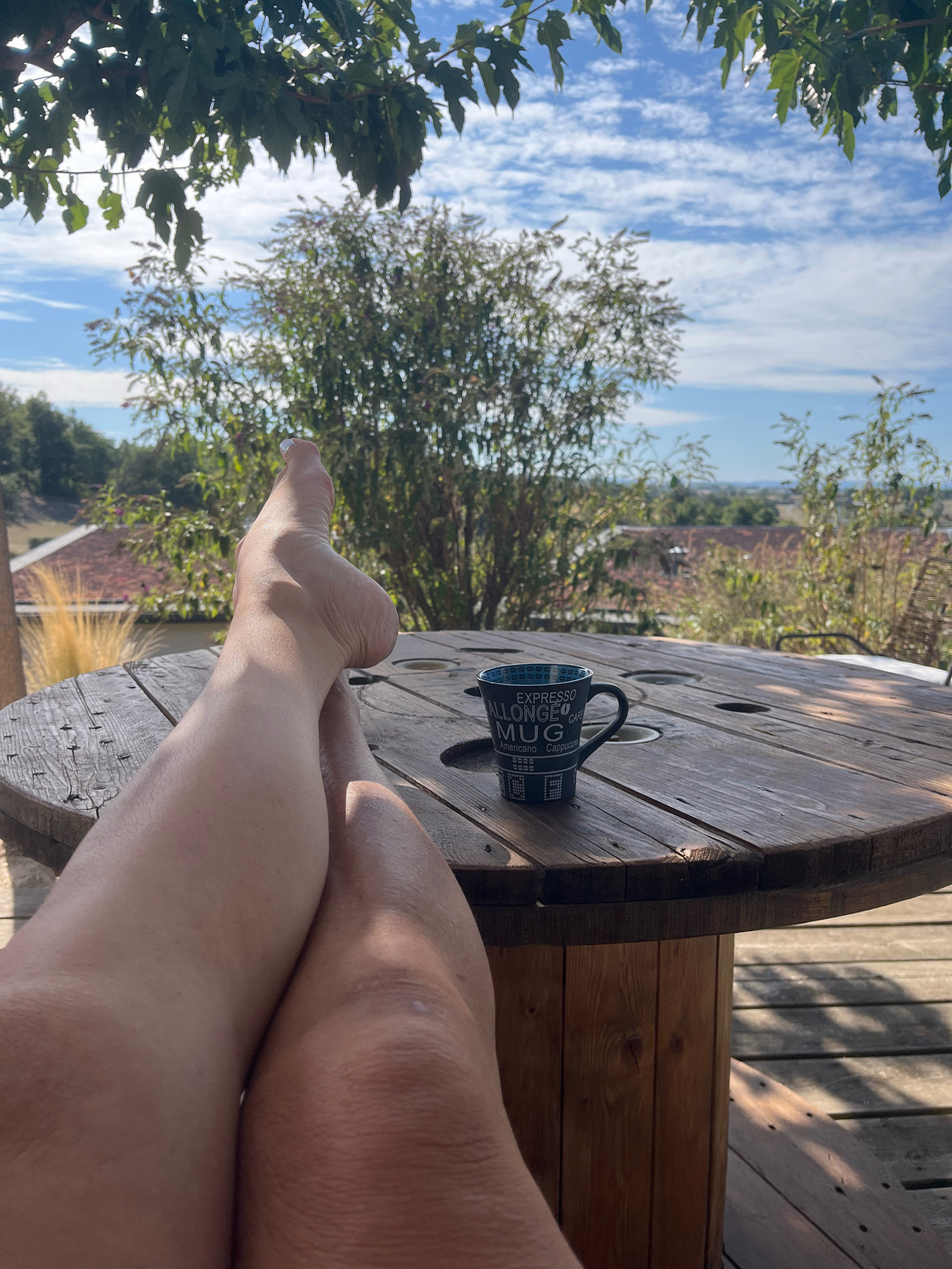 The image depicts a close-up view of a person's bare legs and feet, which are crossed and resting on a wooden table outdoors. A mug with text that reads "EXPRESSO ALLONGE MUG CAFE" is placed on the table near the legs. The table appears to be made from a repurposed wooden cable spool. In the background, lush green trees and foliage are visible against a bright blue sky with scattered white clouds. Beyond the trees, a distant landscape with buildings and rolling hills suggests a rural or semi-rural setting. The scene implies a relaxed, outdoor setting, possibly a patio or deck. The location is identified as Chambœuf, France.