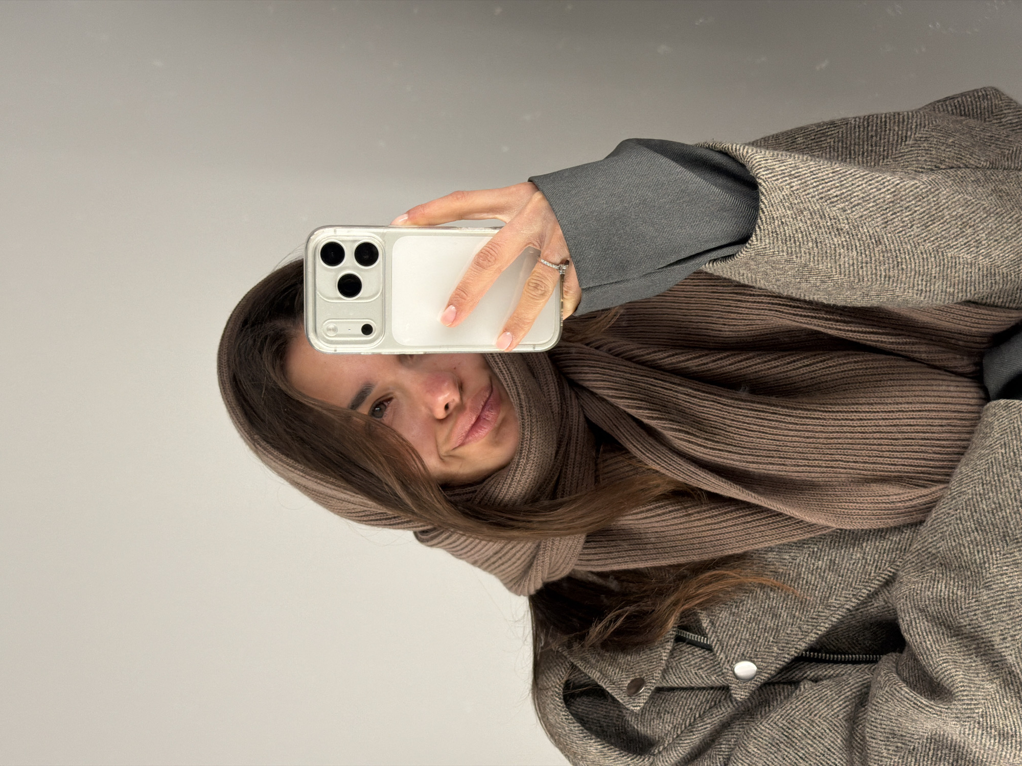 A mirror selfie captures an individual, likely female, from the mid-chest up. The person has long brown hair and is partially obscured by a light brown, ribbed knit scarf wrapped around her head and neck. Only her right eye, nose, and mouth are fully visible, revealing a subtle, neutral expression.
She holds a modern smartphone, identifiable by its triple camera array and clear case, in her right hand, which also displays nails with a light pink French manicure and a sparkling ring on the ring finger.
Her attire consists of a warm, textured coat featuring a herringbone or tweed pattern in grey and beige tones, complete with a visible zipper and snap buttons. A dark grey long-sleeved garment is visible underneath the coat sleeve.
The setting appears to be indoors, against a plain, light grey or off-white surface, suggestive of a wall or mirror. The warm clothing, including the thick scarf and heavy coat, indicates cold weather conditions, aligning with the location context of Krasnoyarsk, Russia. No text is visible in the image. The image itself is presented rotated 90 degrees counter-clockwise.
