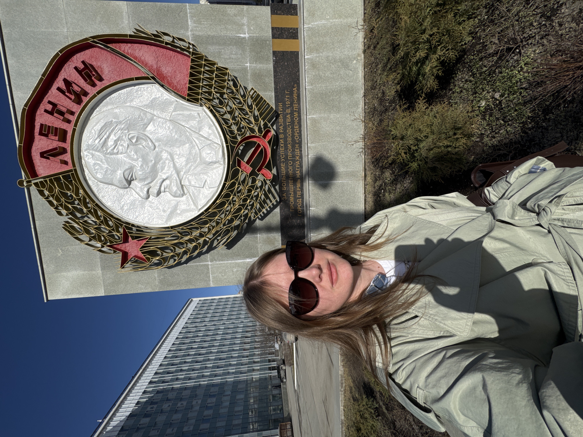 The image presents an outdoor scene in Perm, Russia, featuring a female subject taking a selfie in front of a prominent monument. The subject, a woman with long, light brown hair, wears dark sunglasses and a light-colored (possibly sage green or light khaki) trench coat over a white shirt, looking directly into the camera. Her shadow is distinctly cast across her upper body due to bright sunlight.
The primary background element is a large, grey stone monument, identified as the "Monument to the City of Perm." This monument features a central circular emblem with a white relief bust of Vladimir Lenin in profile, facing left. Above Lenin, the Russian word "ЛЕНИН" (Lenin) is displayed in red, stylized lettering against a red banner. The emblem is framed by a golden wreath design, topped by a red star and featuring a red hammer and sickle symbol at its base. Below the emblem, a black plaque with gold trim is inscribed with Russian text, translating to: "For great successes in the development of industrial production in 1971. The city of Perm was awarded the 'Order of Lenin'."
To the left of the monument, a large, multi-story building with a grid-like facade of numerous windows is visible in the middle ground under a clear, bright blue sky. A paved area, likely a parking lot, with a few vehicles including a dark SUV, separates the monument from this building. The immediate foreground and base of the monument show sparse, low-lying green and brown vegetation. The overall environment suggests a spacious urban area under clear, sunny weather conditions, evidenced by strong shadows.