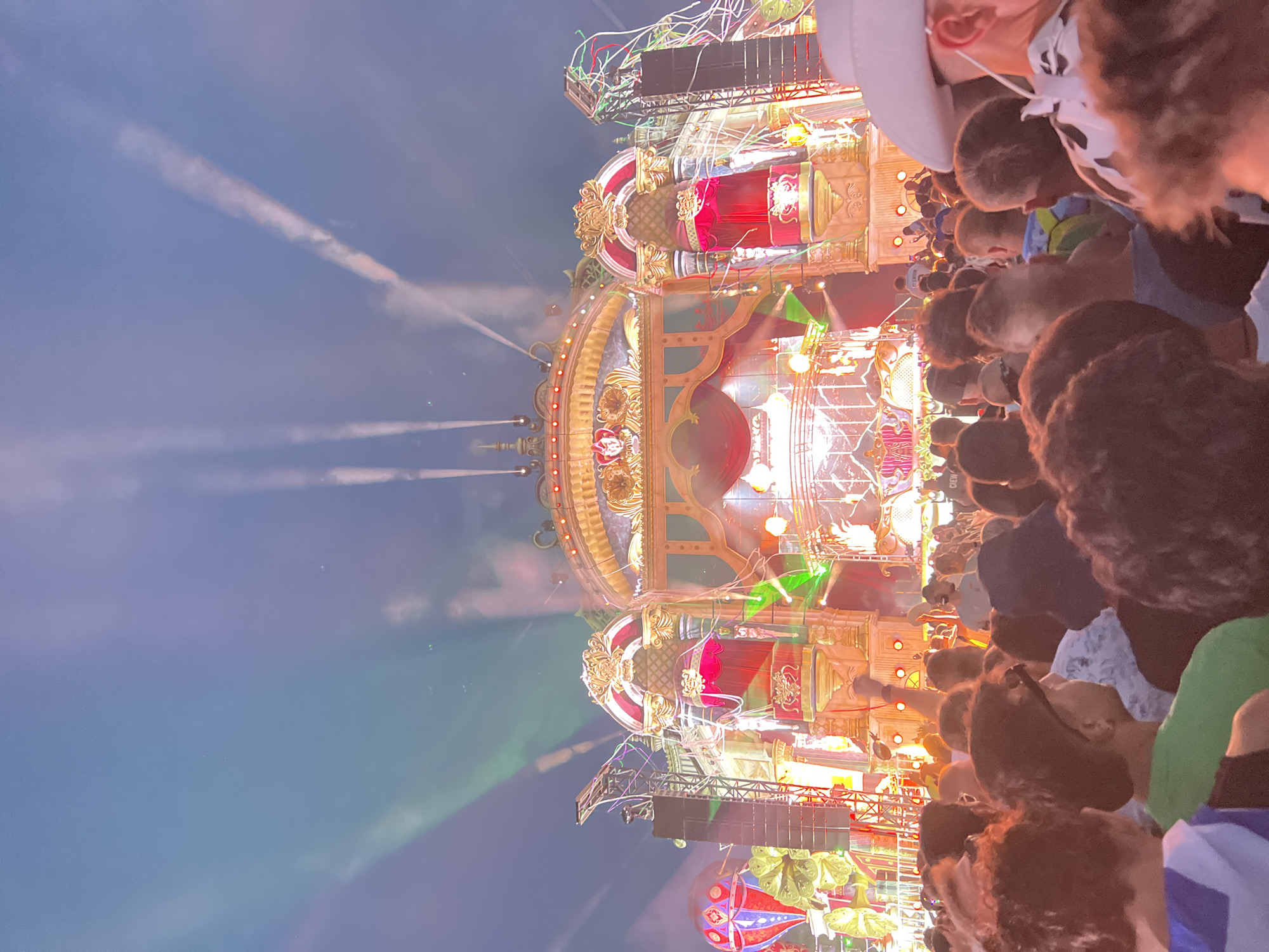 The image depicts a nighttime outdoor music festival scene, specifically at the Tomorrowland festival in Rumst, Belgium. A large, elaborate stage with intricate golden and red decorations dominates the background. The stage features multiple levels, spotlights, and what appears to be a red curtain with golden trim. Smoke or fog effects are visible, illuminated by green and white lights projected from the stage and into the sky, creating beams that extend upwards. In the foreground, a diverse crowd of festival-goers is visible from the back. People are dressed in casual clothing, with some wearing hats and some with their arms raised. The overall atmosphere is one of a vibrant, energetic event.