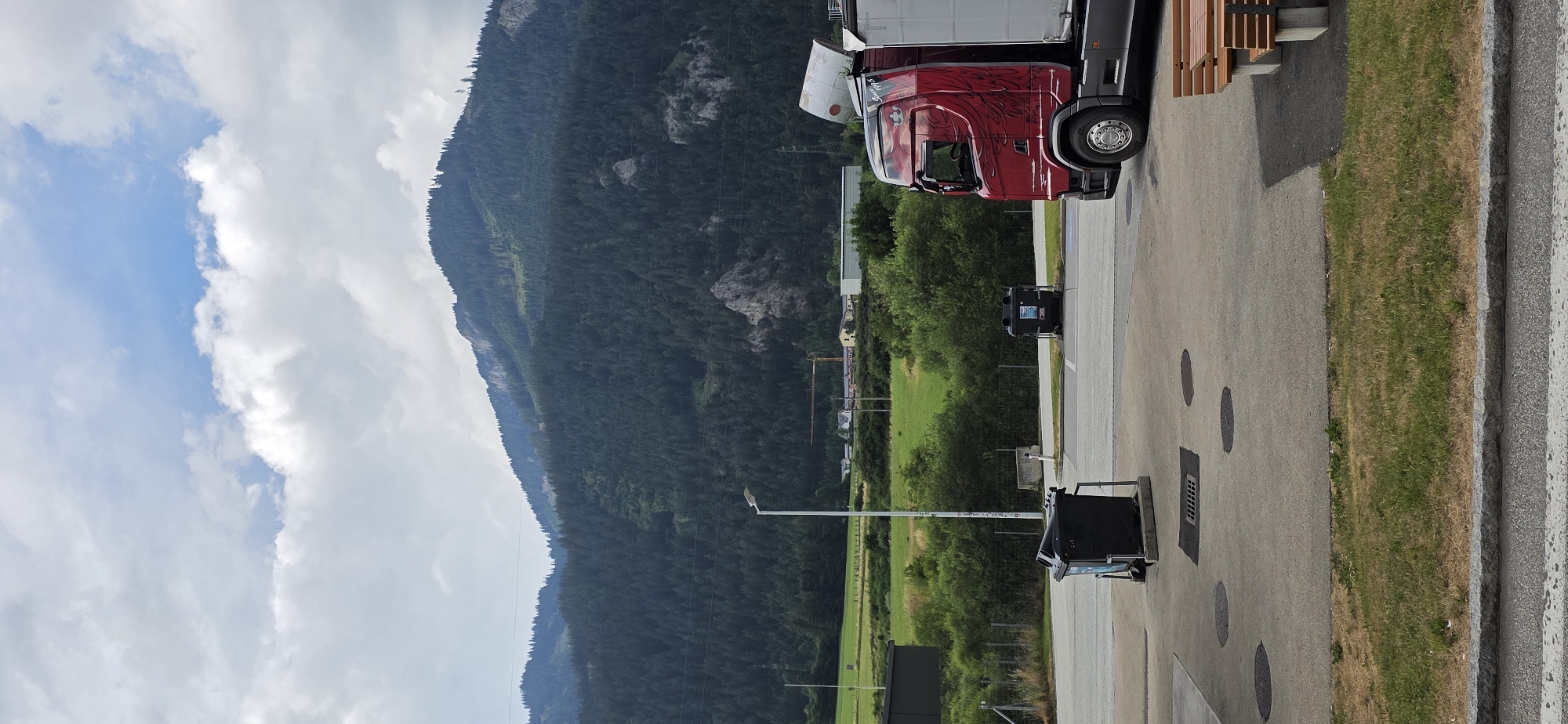 The image depicts a red semi-truck parked in what appears to be a service area or parking lot. The truck has a distinctive graphic on its side and a white cylindrical object protruding from its trailer. In the foreground, two black garbage bins are visible, one with its lid open. The ground is paved with concrete and features several circular manhole covers and a grate. A strip of dry grass with some green patches runs along the edge of the pavement.
Behind the service area, a line of green trees and bushes forms a natural barrier. Further in the background, a vast, densely forested mountain slopes upwards, its dark green foliage contrasting with the lighter green of the fields visible in the mid-ground. The sky is partially cloudy with patches of blue visible. In the distance, a crane and some buildings suggest a small town or industrial area nestled in the valley.
The scene is likely located in Kammern im Liesingtal, Austria, given the mountainous terrain and typical European architectural style suggested by the distant buildings. The overall impression is one of a rural or semi-rural setting, possibly near a highway rest stop or a logistics hub, with the imposing natural landscape dominating the background. There are no people visible in the frame.