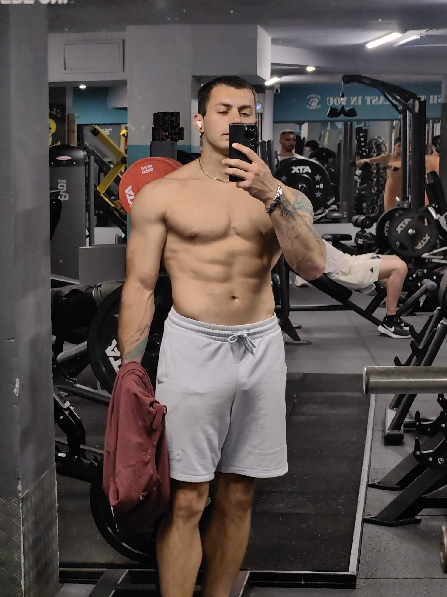 A man is taking a mirror selfie in a gym located in Varna, Bulgaria. He is shirtless and wearing light gray athletic shorts. He holds a dark-colored smartphone in his right hand, which he uses to take the photo. He is also holding a dark red t-shirt in his left hand. The man has tattoos on his left arm and is wearing white wireless earbuds and a silver chain necklace. In the background, other gym equipment and people are visible. The gym appears to be well-equipped with various weight machines and free weights. The overall lighting is typical of an indoor gym environment.