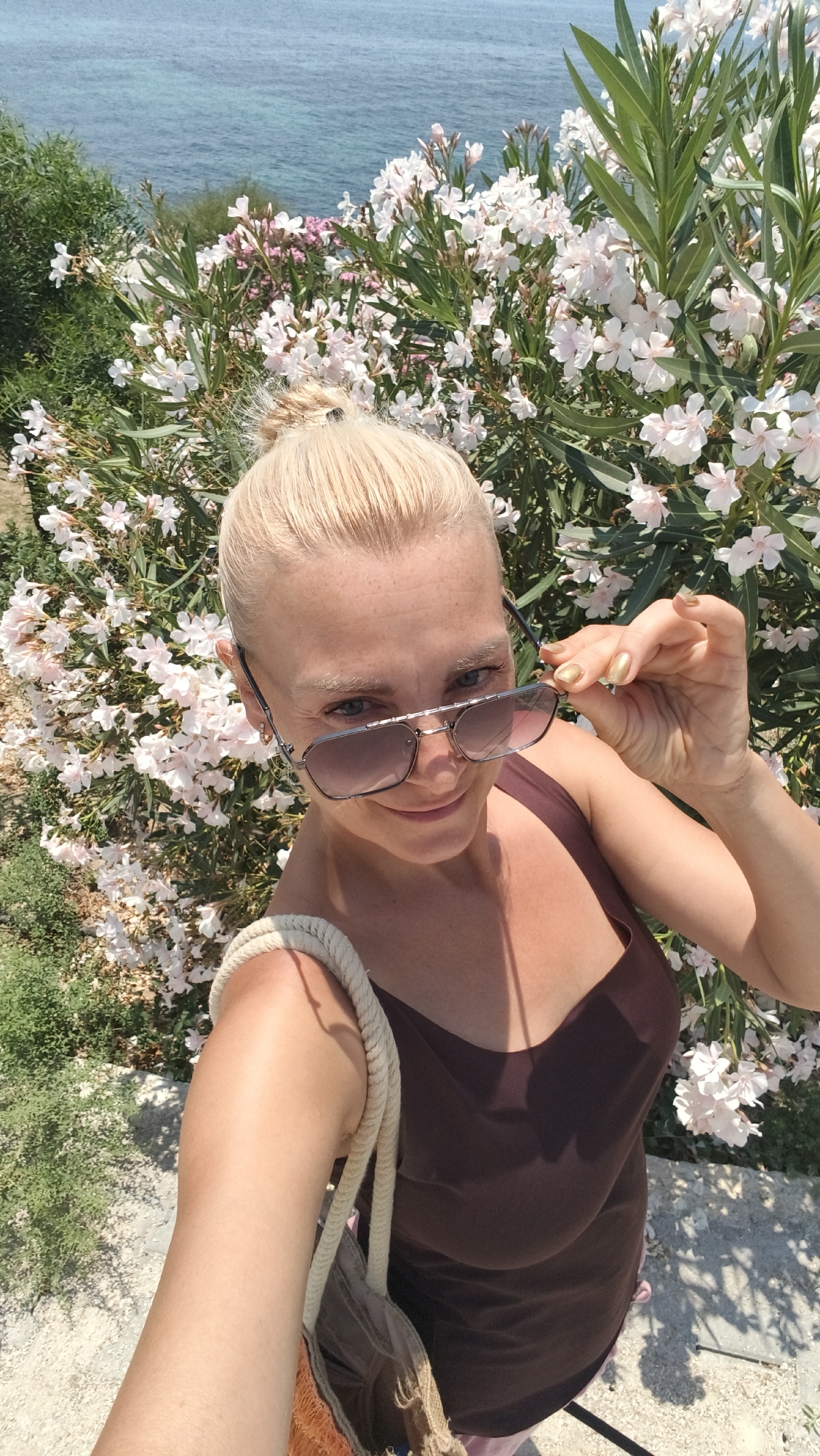 In Kuşadası, Turkey, a blonde woman with her hair in a bun takes a selfie in front of a bush with white flowers. She is wearing sunglasses and a brown tank top, and a straw bag hangs from her shoulder. The ocean is visible in the background.