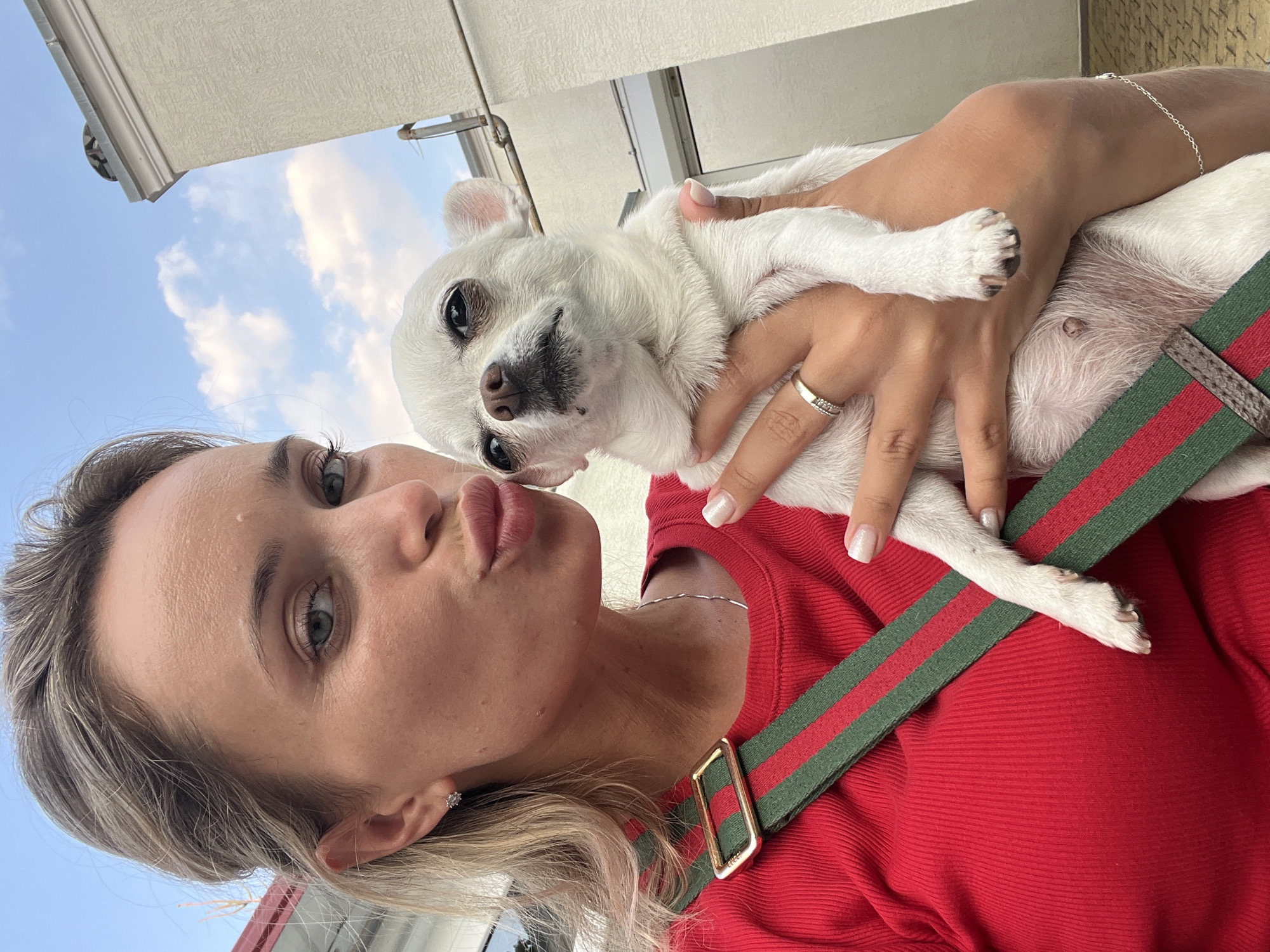 A young woman with blonde hair pulled back and light blue eyes is captured in a selfie, holding a small white chihuahua. She is wearing a red ribbed top with a green and red striped strap. The dog, with its ears perked and a somewhat disgruntled expression, is held in her arms. A ring is visible on the woman's left hand, and she appears to be puckering her lips towards the dog. The background shows a building and a cloudy blue sky. The scene takes place in Fontanka, Ukraine.