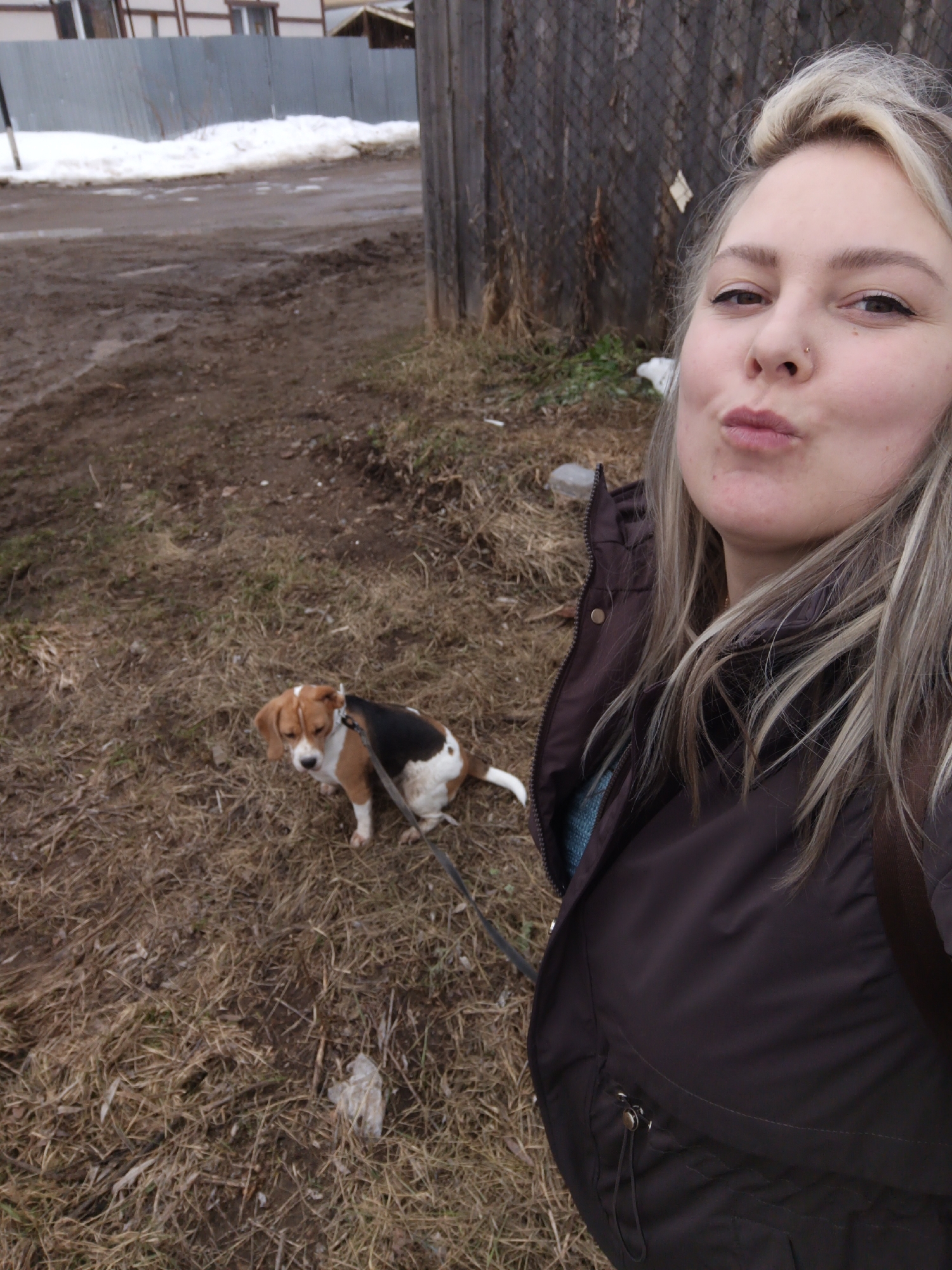 The image is a selfie taken outdoors in what appears to be a rural or suburban area. A woman with blonde hair and a nose ring is in the foreground, making a kissing face towards the camera. She is wearing a dark brown jacket. Beside her, on the left, is a small beagle dog sitting on dry grass. The dog is leashed, and the leash extends towards the lower right of the frame.
The background shows a muddy area with patches of snow and some buildings and a wooden fence in the distance. Based on the location information provided, this image was taken in Chaikovsky, Russia.