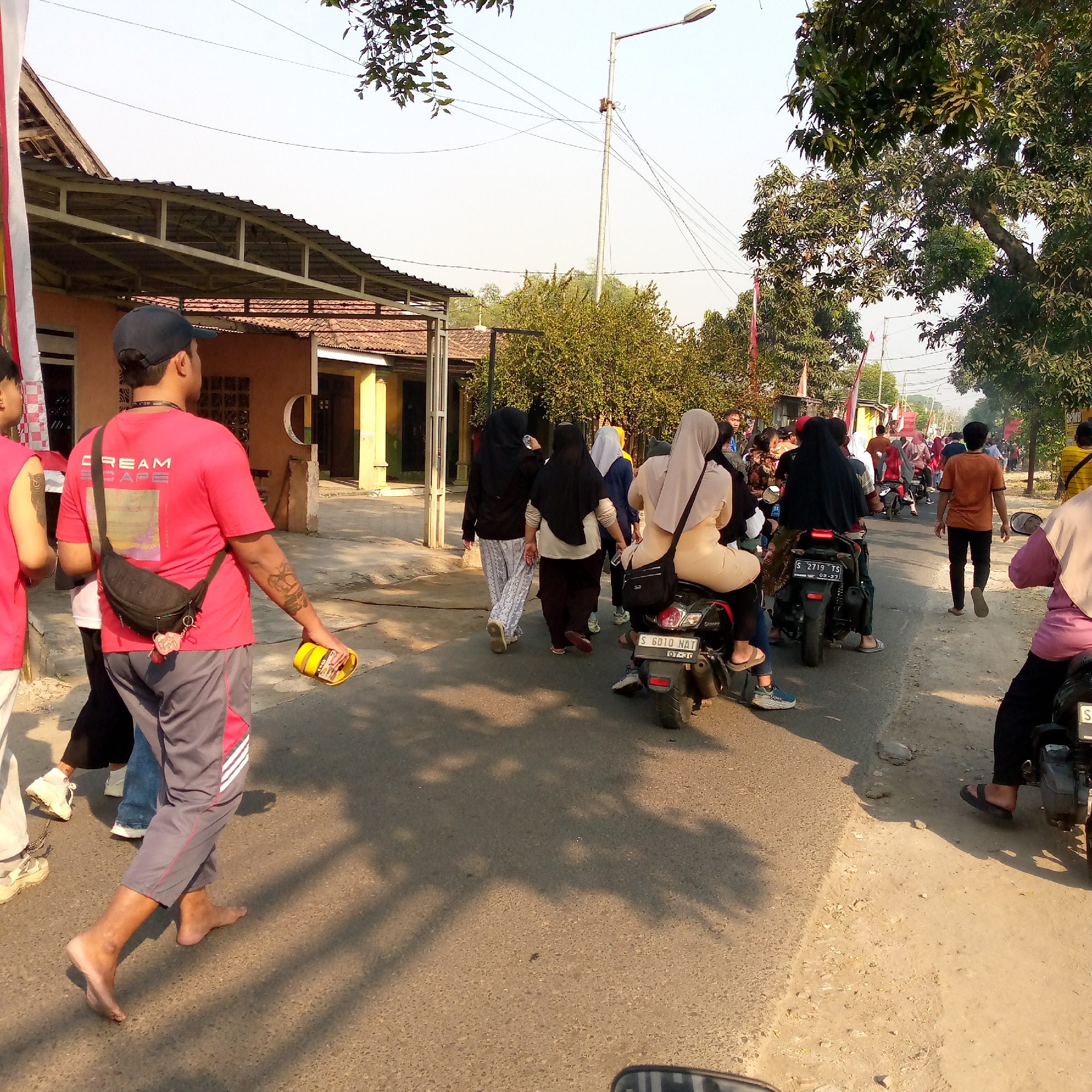 The image depicts a street scene in Tambakagung, Indonesia, under a clear sky. A group of people are walking and riding motorcycles along a paved road. In the foreground, a man wearing a red t-shirt and gray pants with red trim is walking barefoot, carrying a black sling bag and holding a yellow object in his right hand. Several other individuals are seen walking ahead of him, some wearing headscarves. A few people are riding motorcycles, with one motorcycle in the middle ground displaying the license plate "S 6010 NAT". The environment features buildings with tiled roofs, trees, and utility poles with electrical wires. Red and white flags are visible, suggesting a possible cultural or celebratory event. The ground is a mix of pavement and dirt. The overall scene appears to be a busy, casual gathering or procession.