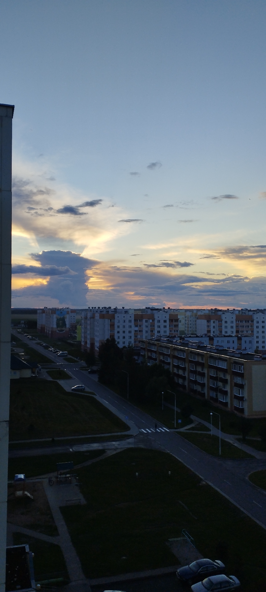 An aerial vertical view captures a twilight scene over an urban area, likely Skidzieĺ, Belarus. The sky is a gradient of pale blue to a soft orange and yellow near the horizon, with dramatic clouds illuminated by the setting sun. In the foreground, the edge of a light-colored building occupies the left side of the frame. Below, rows of multi-story apartment buildings stretch across the landscape, characterized by light-colored facades with orange and yellow accents, and numerous windows. Streets with faint lane markings run between the buildings and through green areas. Several parked cars are visible on the roads and in parking lots. A children's playground structure is discernible in the lower left quadrant. The overall atmosphere is that of a residential neighborhood at dusk.