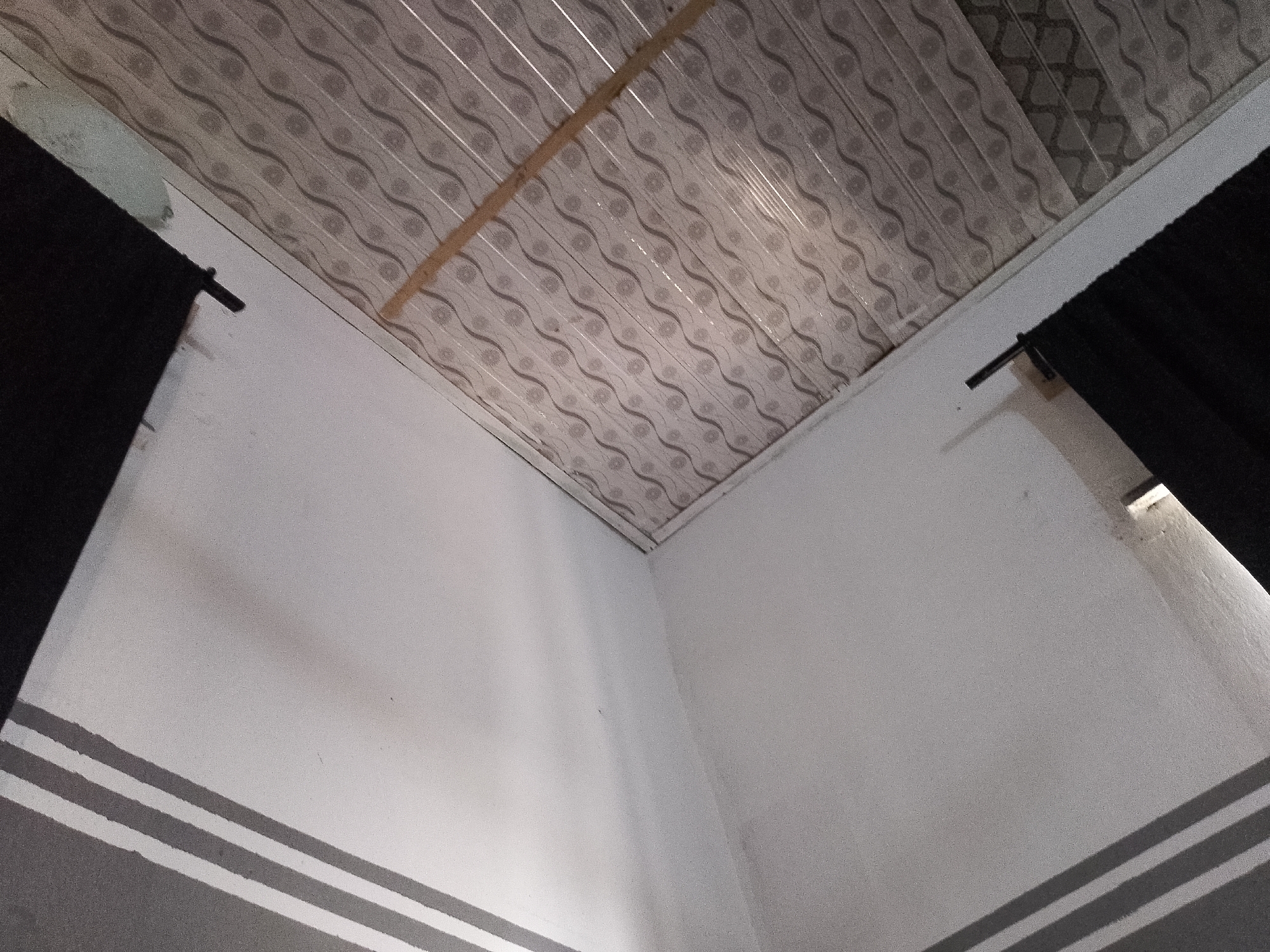 The image shows an interior view of a corner where two walls meet a patterned ceiling. The walls are painted white and have three horizontal grey stripes near the bottom. On the left side, a dark material, possibly a curtain or fabric, is hanging. A black rod or bracket is visible near the top of this hanging material, attached to the wall. On the right side, a similar dark material is also present. The ceiling is made of panels with a repeating wavy pattern and some floral motifs. A light-colored wooden beam runs diagonally across the ceiling. The lighting in the scene appears to be coming from the right, casting subtle shadows on the white walls. There are no people or discernible actions taking place in the image. This scene is consistent with an indoor residential or commercial space in Jauro Yinu, Nigeria.