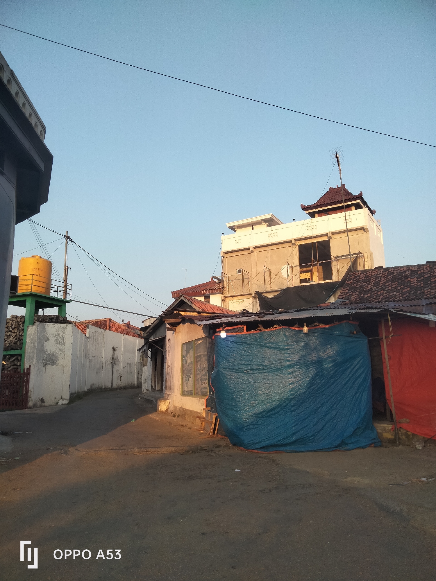 The image shows a street scene in what appears to be a village or a less developed area of a city. In the foreground, the paved road is partially illuminated by sunlight, casting long shadows. The left side of the image features a white wall, behind which a yellow water tank on stilts is visible, with a stack of wood or logs beneath it. A dark fence borders the left edge of the image.
To the right of the road, there are several structures. One building has a dark, corrugated metal roof and is partially covered by a large blue tarp. Adjacent to this is another structure covered by a red tarp. Further back, a multi-story building is under construction, with scaffolding surrounding its upper levels. This building has a distinct, multi-tiered roof, reminiscent of traditional Indonesian architecture, with a dark tile finish and an antenna on top. The sky is clear and light blue. The "OPPO A53" watermark is present in the bottom left corner, indicating the device used to capture the image. The scene depicts a street with residential or semi-commercial buildings, some appearing older and in disrepair, while another is undergoing construction. There are no people visible in the image. The overall impression is that of an ordinary day in a working-class neighborhood.