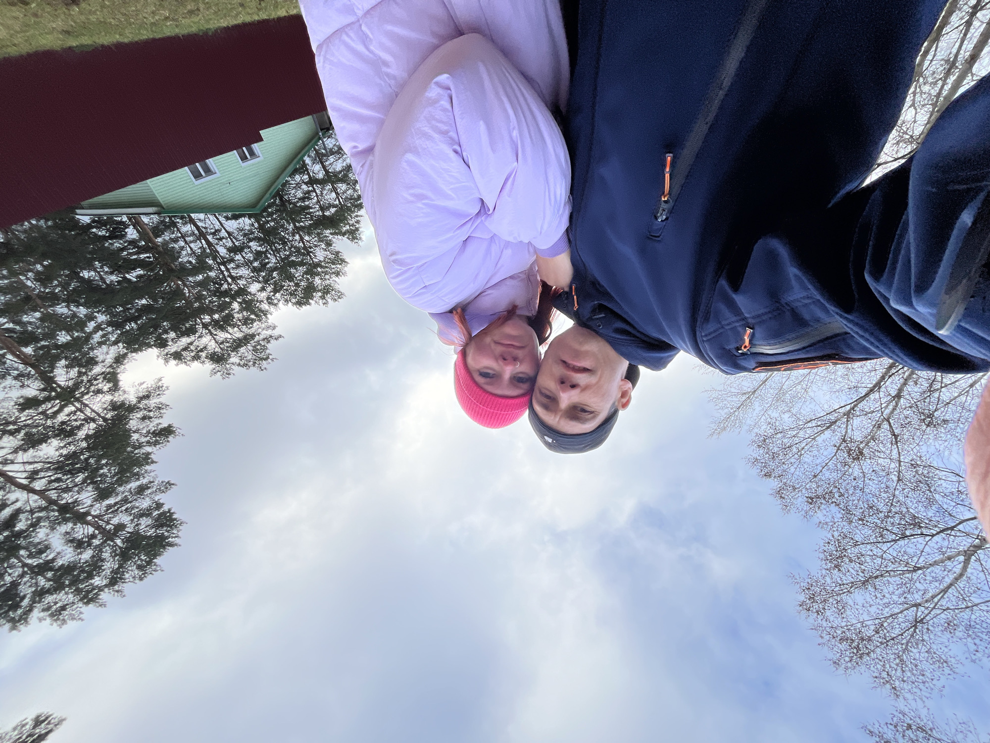 A low-angle selfie captures two individuals, a man and a woman, looking down towards the camera against a backdrop of sky, trees, and a house. The image is taken from a perspective pointing upwards.
The woman is on the left, wearing a light purple puffy jacket and a bright pink knitted beanie. She has reddish-brown hair and a visible nose piercing. The man is on the right, dressed in a dark blue zip-up jacket with orange zipper pulls and a dark-colored beanie, which has a small, light-colored logo on the front that is not clearly legible. Both subjects have neutral to slightly smiling expressions as they look directly into the camera lens.
Behind the individuals, the sky is predominantly visible, showing a mix of light blue and white clouds, indicating an overcast or partly cloudy daytime. To the left side of the background, a light green house with white-framed windows and a section of a dark red roof is partially visible. Tall evergreen trees with dark foliage are prominent in the left and center background, while bare deciduous trees with thin branches are seen in the right background.
The clothing suggests cool weather, possibly late autumn or early spring. The overall lighting is soft and diffused, consistent with a cloudy daytime setting. The location is Chaikovsky, Russia.