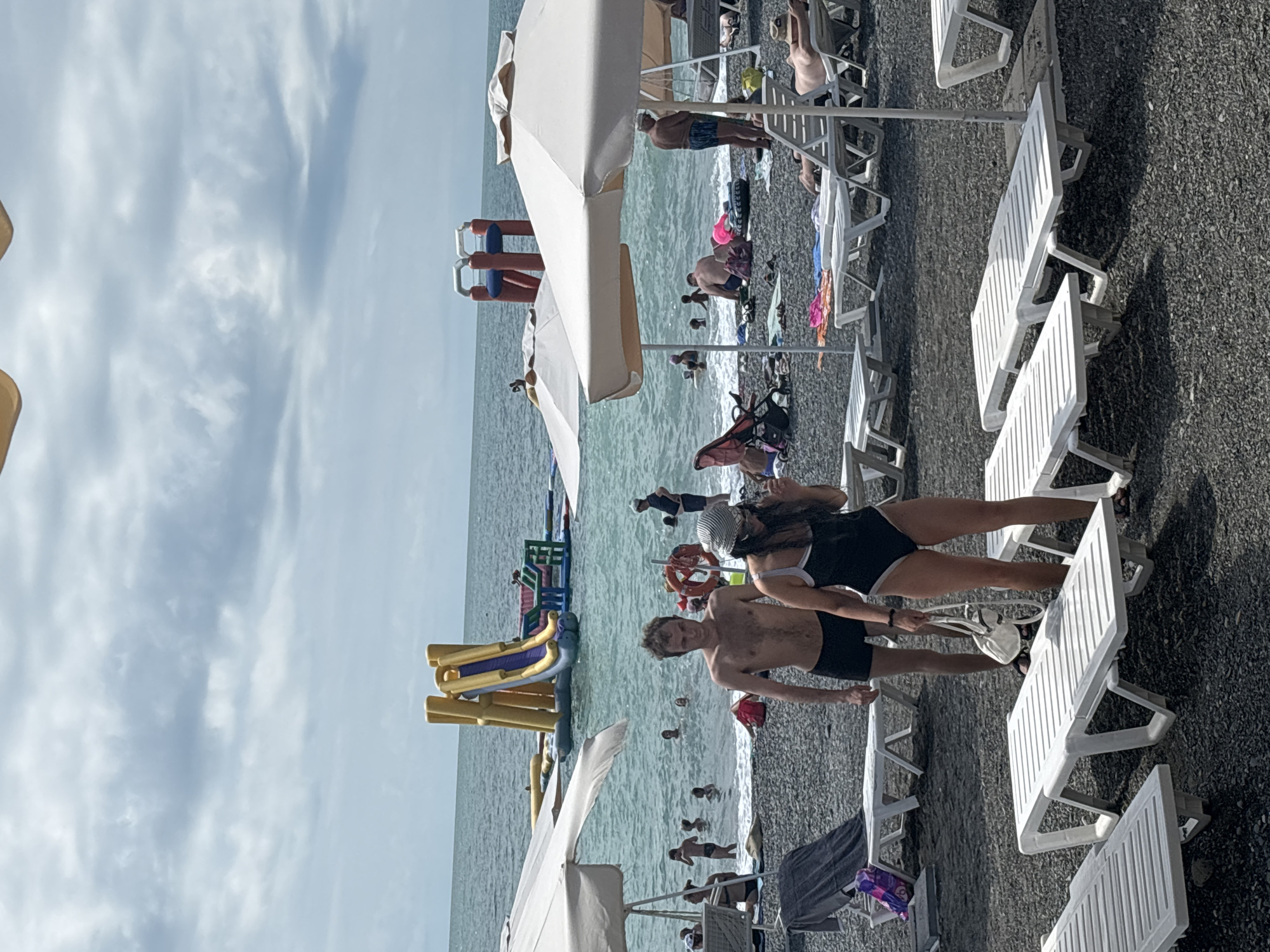 The image depicts a crowded beach scene, likely in Sochi, Russia, given the context of the provided crops and the common beach resorts in the area. In the foreground, several empty white plastic lounge chairs are arranged on a dark pebble beach. A man and a woman, both dressed in swimwear, are walking away from the camera towards the water. The woman is wearing a black and white swimsuit and a hat, and carrying a white bag. The man is shirtless and wearing black swim trunks.
In the midground, the sea is populated with many people swimming and enjoying various inflatable attractions. Prominently visible is a large yellow and purple inflatable water slide. Further back, other inflatable structures and a small red inflatable can be seen on the water. Beach umbrellas provide shade for some of the beachgoers. The sky is overcast with clouds. The overall atmosphere suggests a busy summer day at a popular beach resort.