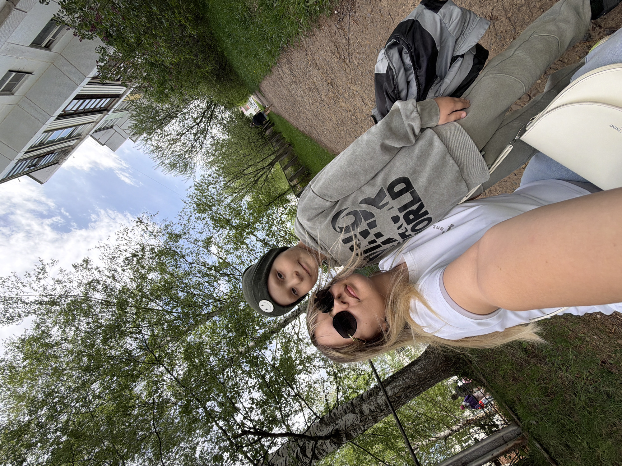 The image shows two individuals, likely a mother and child, posing for a selfie. The woman has blonde hair and is wearing sunglasses and a white shirt. The child is wearing a gray hoodie and a green beanie with a white circular emblem. They are standing on a path, with a building visible on the left side of the frame and trees surrounding them. Based on visual markers, the location is the city of Chaikovsky, Russia. The background includes trees and a clear sky with clouds. The path is made of gravel. A gray jacket and a white purse are visible near them. They are interacting by posing for a photo together.