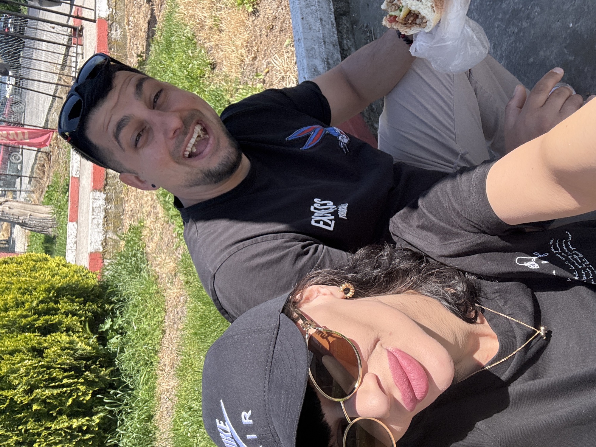 A man and a woman are posing for a selfie in an outdoor setting. The man is on the left, wearing a black t-shirt with an image of Bugs Bunny, sunglasses on his head, and he is holding a sandwich in a plastic bag. He is smiling broadly, exposing his teeth. The woman is on the right, wearing a black t-shirt, a black Nike cap, and sunglasses. She is wearing pink lipstick and a gold necklace. They are sitting on a grey concrete surface with red and white painted edging. In the background, there is grass and a bush on the left, and a grassy area on the right. There are cars and buildings visible in the distance, indicating an urban or suburban location. This is in Probuda, Bulgaria.
