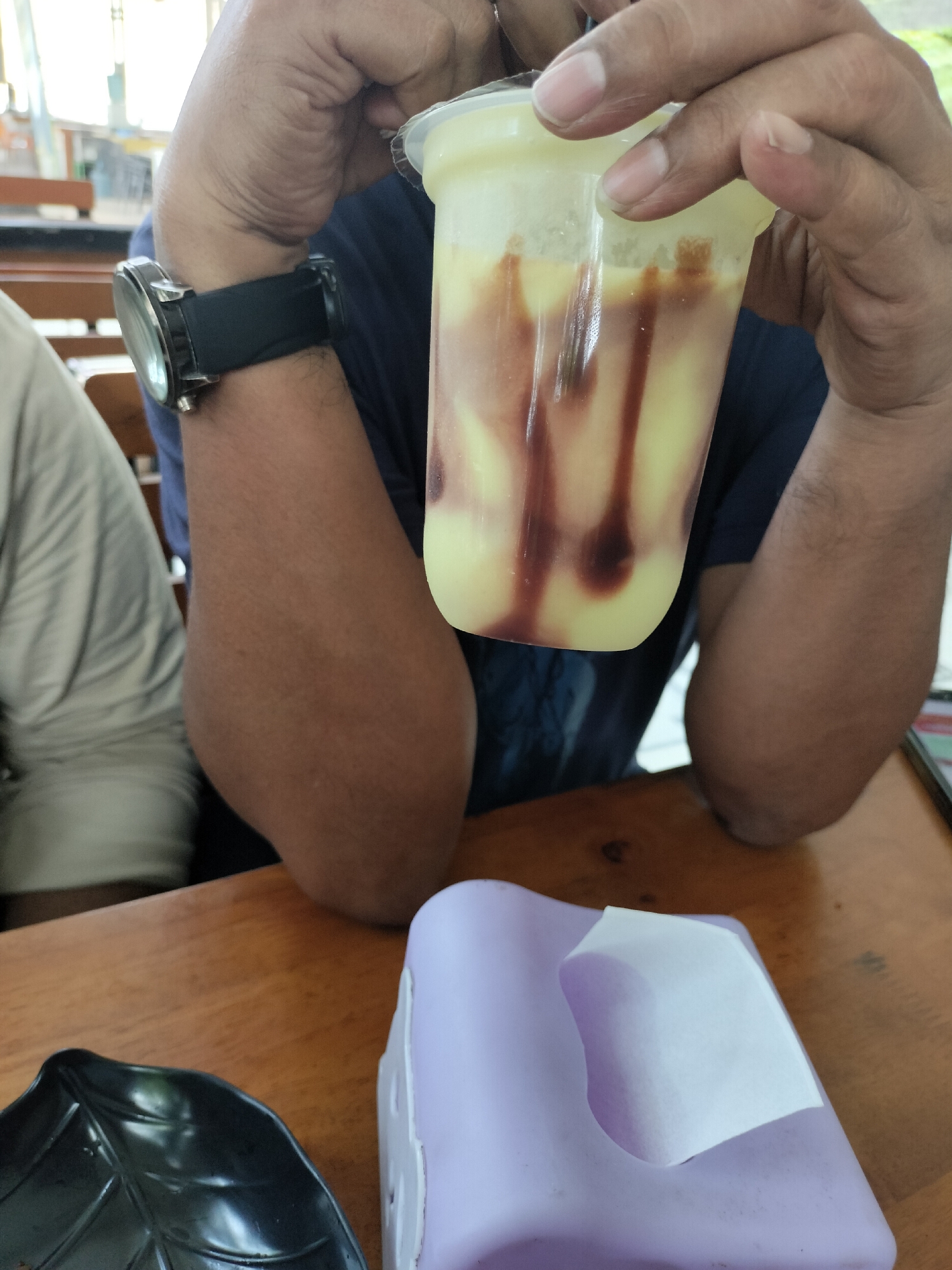 The image captures a close-up of a person holding a plastic cup containing a light yellow beverage with dark streaks, likely a drink with a sauce or syrup. The person is wearing a dark t-shirt and a black wristwatch on their left arm. Another person, wearing a light-colored shirt, is partially visible on the left side of the frame. In the foreground, a wooden table holds a black leaf-shaped dish on the left and a purple tissue dispenser with a white tissue on the right. The background suggests an indoor setting with wooden furniture and windows, possibly a cafe or restaurant in Keudealue Ie Puteh, Indonesia.