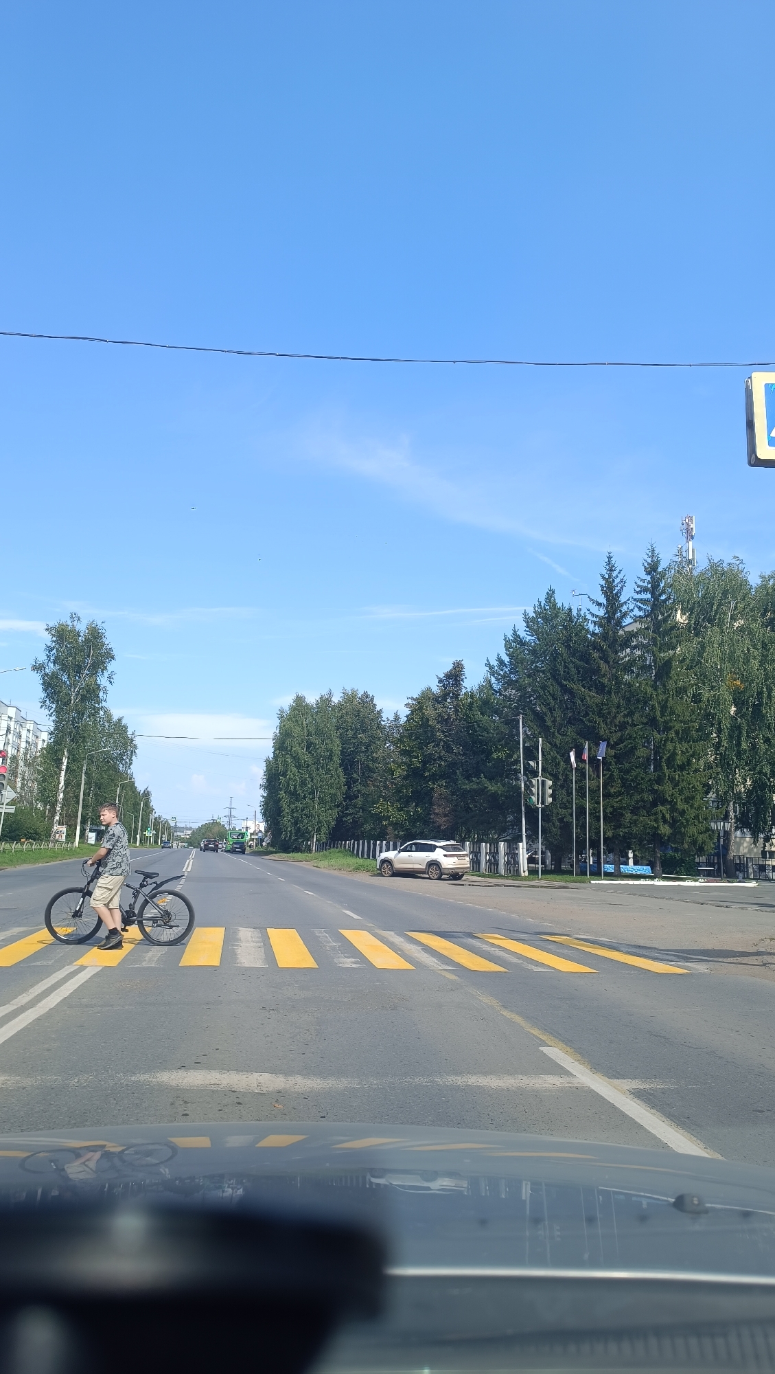 A young man rides a bicycle across a marked pedestrian crossing on a sunny day in Chaikovsky, Russia. The road has yellow striped markings for the pedestrian crossing, and there are lane markings. To the left, there are apartment buildings and trees. To the right, there are more trees and a white SUV parked on the side of the road. In the distance, there are other vehicles on the road and cell towers. The sky is clear and blue with a few wispy clouds. The image is taken from the perspective of someone inside a vehicle, as indicated by the reflection on the dashboard in the foreground.