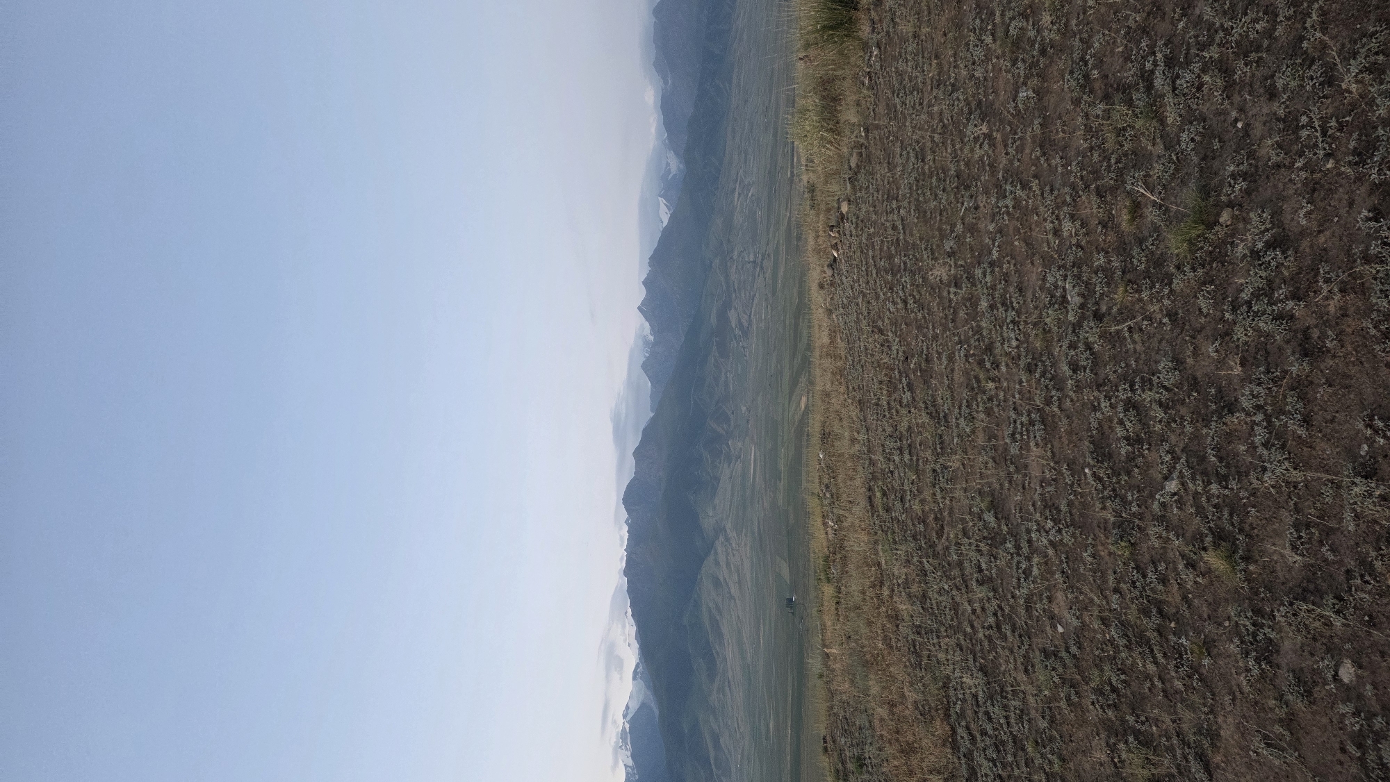 The image captures a panoramic view of a mountainous landscape. In the foreground, dry, sparse vegetation covers the earthy ground. A gentle slope leads down to a vast, flat valley dotted with fields, suggesting agricultural activity. In the mid-ground, a distant structure, possibly a house or farm building, is visible. The background is dominated by a majestic mountain range, with snow-capped peaks visible in the distance. The sky above is a clear, pale blue, transitioning to a lighter hue near the horizon, indicating either early morning or late evening light. The overall atmosphere is serene and natural, characteristic of a rural or undeveloped area. The location is identified as Ак Жар, Kyrgyzstan. There are no people or discernible actions depicted in the scene.