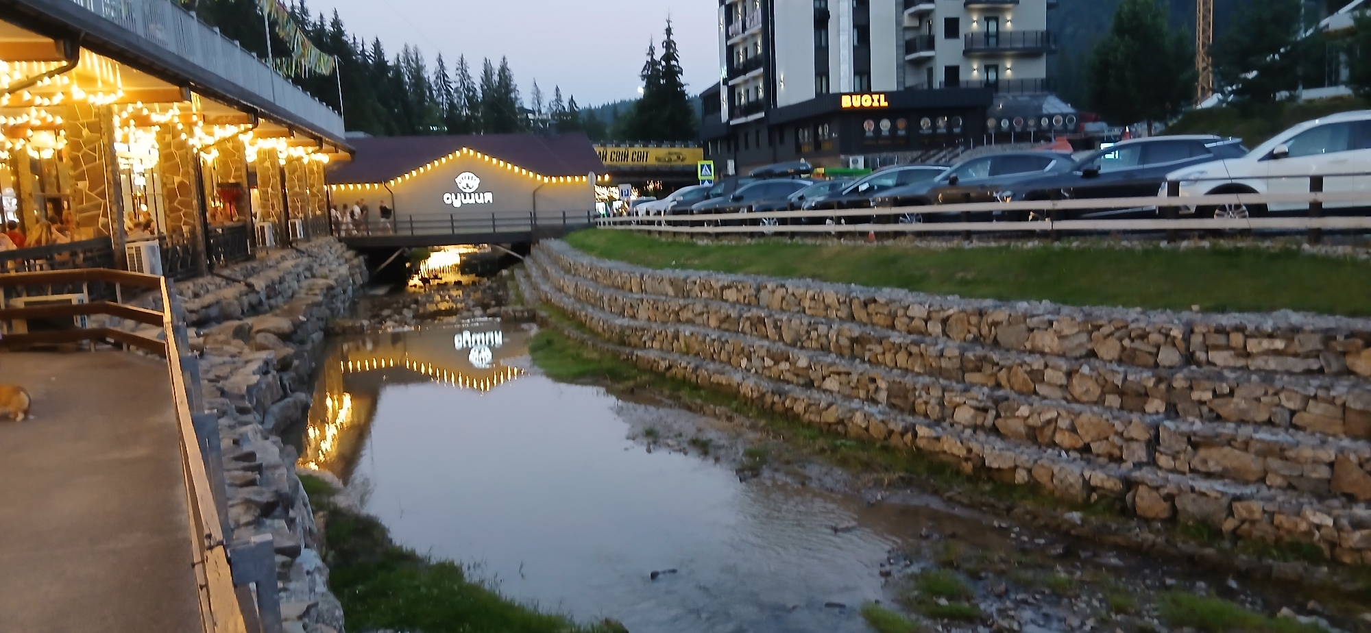 The image captures a scene in an outdoor, possibly resort, setting. In the foreground, a stone-lined stream or canal flows. The canal banks are reinforced with stacked stone walls. Beyond the canal, a bridge crosses over it, leading to a building with illuminated signage that reads "ОЛІМП". This building is decorated with string lights. To the left of the canal, a stone building with an outdoor seating area is visible, also adorned with numerous string lights, suggesting it is a restaurant or cafe. A small dog is visible on the paved area in front of this building. Further in the background, to the right of the "ОЛІМП" building, a multi-story modern building stands, with the word "BUGOL" illuminated on its facade. A row of parked cars is visible behind a white fence in front of the modern building. The scene appears to be at dusk or early evening, indicated by the low light and the illuminated lights. Pine trees and a hilly landscape are visible in the background.