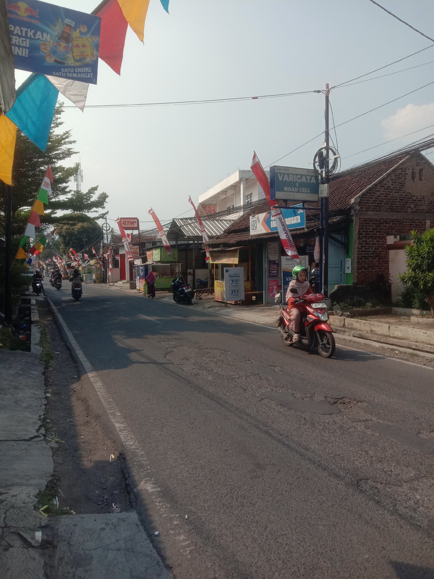 This image depicts a street scene in Cianjur, Indonesia, characterized by its bustling atmosphere and festive decorations. The street is paved with asphalt and shows signs of wear, with several potholes visible. The sky is clear and bright, indicating a sunny day. On the left side of the road, a sidewalk is visible, bordered by a drainage ditch.
The street is lined with various small businesses and shops, many of which are adorned with colorful flags and banners. Some banners feature advertisements, including one for Red Bull. Indonesian flags, both small and large, are prominently displayed, suggesting a national celebration or event. The buildings along the street vary in architectural style, with some appearing to be single-story shops and others with multiple stories. One building displays a sign for "VARIGATA X NOAH CELL," indicating a telecommunications or electronics store.
Several individuals are present in the scene, primarily riding motorbikes. A woman in a light orange outfit and green helmet is riding a red motorbike in the foreground, heading towards the viewer. Other motorbikes are seen further down the street, with riders either moving or parked. A person in a pink hijab is standing near a storefront on the right side of the road. Trees and vegetation are visible, adding a touch of greenery to the urban environment. The overall impression is one of a lively local street in Indonesia, likely during a period of celebration.