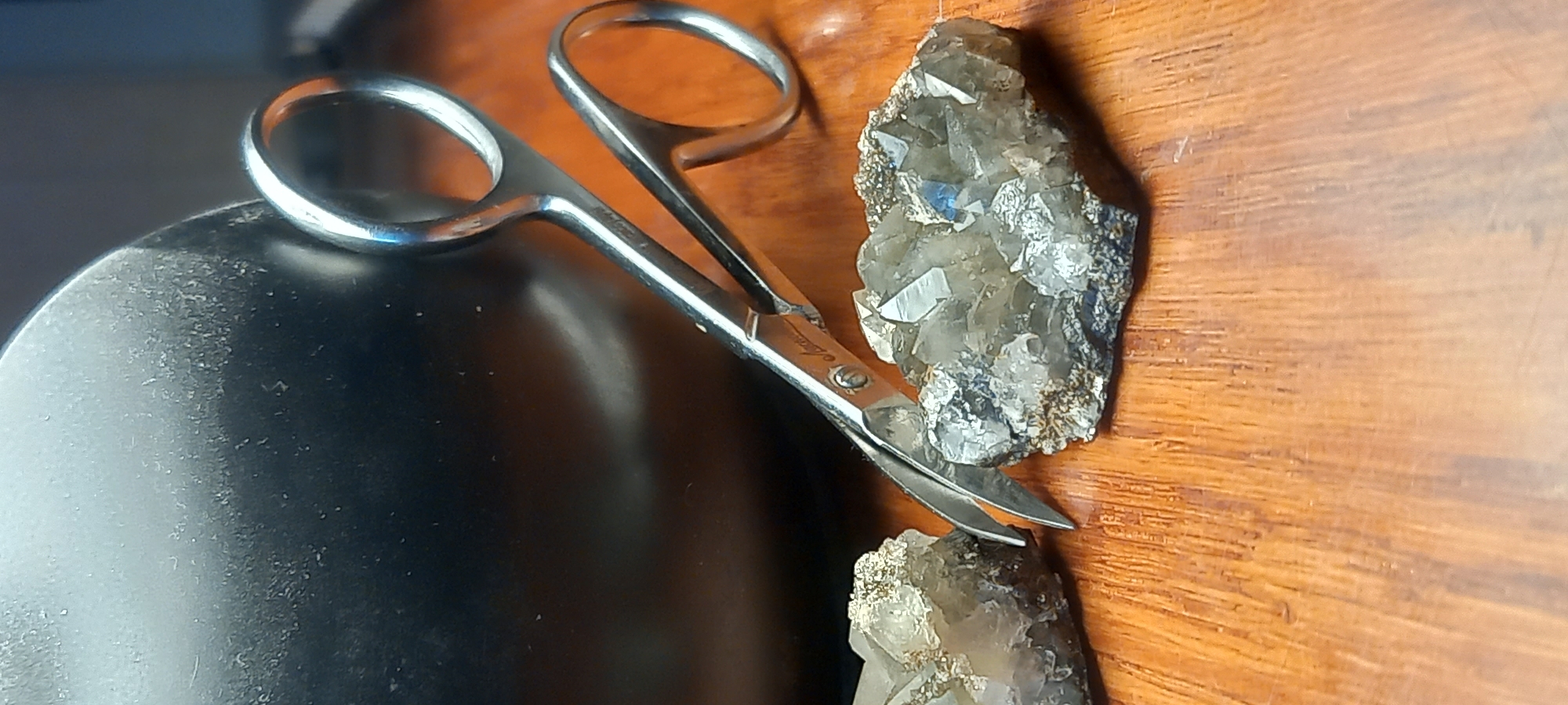 The image presents a still life composition featuring a pair of silver-colored manicure scissors, two mineral specimens, and two distinct background surfaces. The scissors are positioned diagonally across the frame, bridging a dark, curved object on the left, possibly a metal or plastic bowl, and a wooden surface on the right. Visible text on the side of the scissors includes "Stainless" and what appears to be "Ostrov."
Two pieces of mineral, likely quartz, are present. A larger specimen occupies the upper right portion of the wooden surface, showcasing numerous small, translucent, and grayish crystals on a rock matrix, with a hint of blue within one crystal cluster. A smaller, similar mineral piece is situated in the lower right, with the tips of the scissors hovering above it. The wooden surface displays a warm, light brown tone with visible grain patterns.
The lighting highlights the reflective surfaces of the scissors and the dark object, creating subtle bluish reflections on the metal. The scene is static, depicting no activity or individuals, and appears to be set indoors, possibly on a table or workbench. There are no explicit visual cues within the image to indicate time of day, weather, or specific geographic location like Kryvyi Rih.