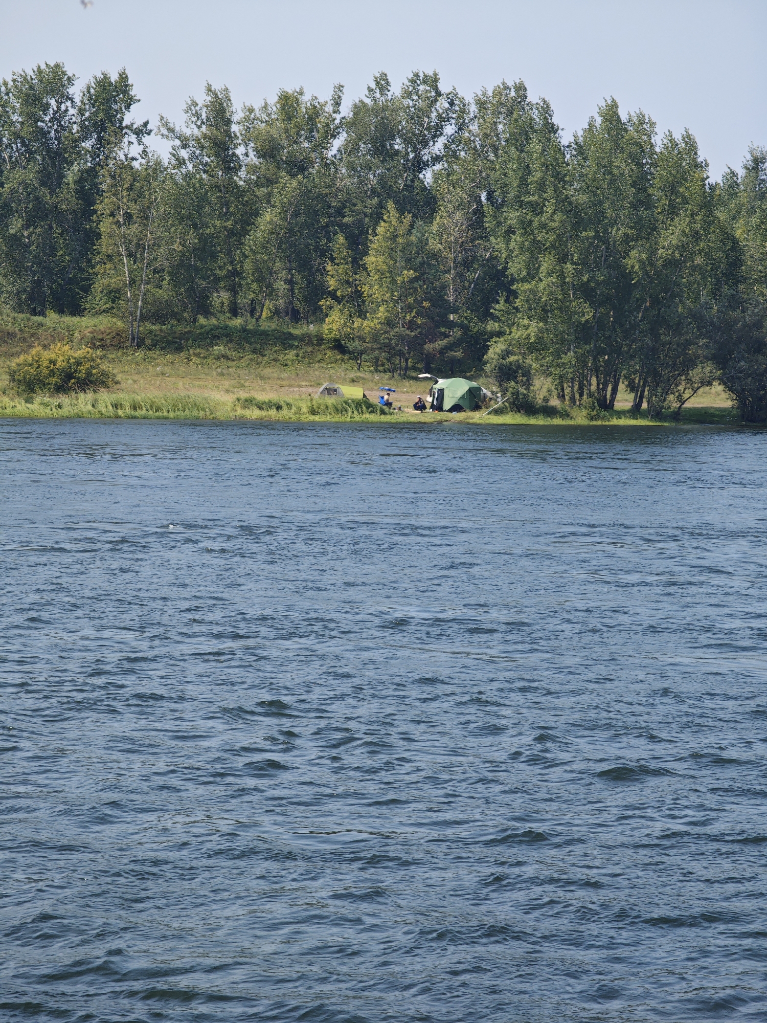 The image depicts a wide, blue river with moderate ripples under a clear, pale blue sky. In the background, a lush green treeline forms the distant bank. Along the edge of the river, a grassy area is visible, with a small camping setup. A green tent is pitched, and several people are seated near it. The setting appears to be a natural outdoor area, likely a park or recreational zone along the Yenisei River within the city of Krasnoyarsk, Russia. The overall scene conveys a peaceful, summery atmosphere.