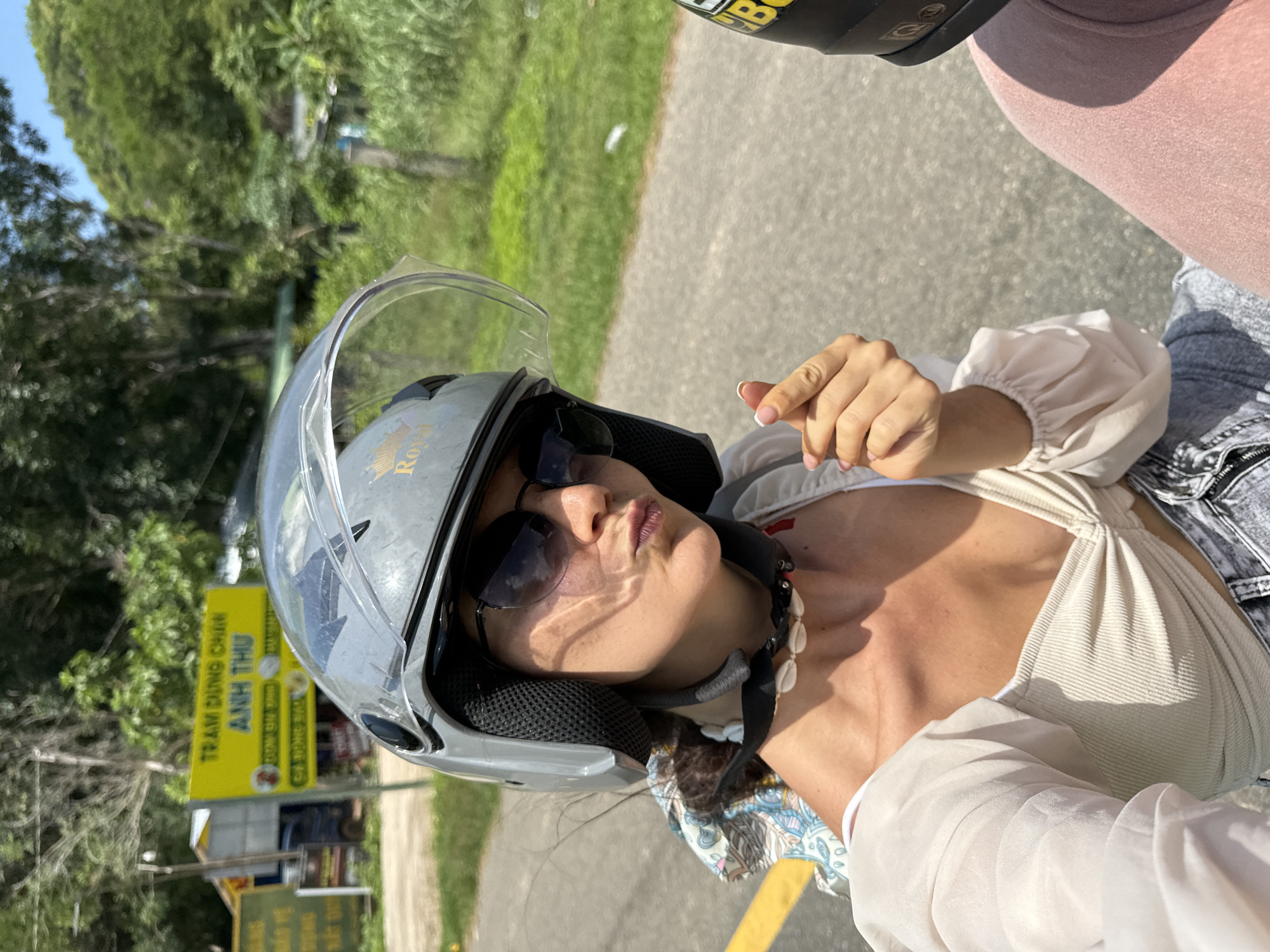 In Ham Thuan Bac, Vietnam, a woman wearing a grey helmet and sunglasses is riding on a motorcycle. She is looking at the camera and making a gesture with her fingers. The background shows a street with trees and buildings.