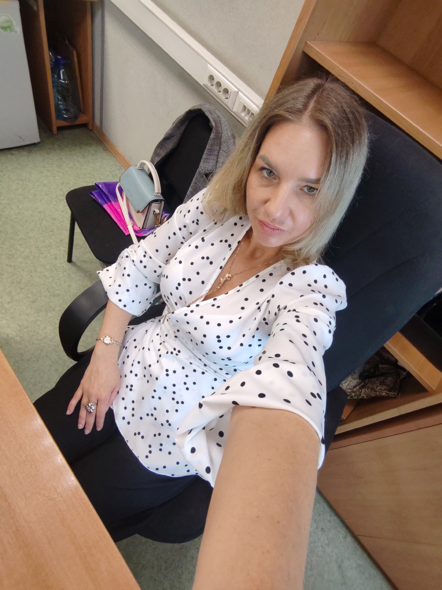 A blonde woman is captured in a selfie, seated in a black office chair within what appears to be an office or workspace. She has light-colored eyes and a direct, neutral gaze towards the camera, holding the device with her extended right arm. She is wearing a white blouse adorned with black polka dots, black pants, a gold necklace with a pendant, a gold-toned watch on her left wrist, and a prominent ring on her left ring finger.
The background reveals a functional environment. To her left, a white appliance (possibly a refrigerator) stands next to a wooden shelving unit holding a large blue water bottle. A light-colored wall features a white electrical outlet strip with multiple sockets. To her right, more wooden shelving or cabinetry is visible. Another black office chair is positioned behind her, draped with a gray jacket, and holds a light blue purse along with some colorful folders or bags. A wooden desk surface is partially visible in the foreground on both sides of the image. The floor is covered with a greenish-grey patterned carpet or linoleum. The lighting suggests an indoor setting, likely relying on artificial sources. There is no discernible text visible in the image.