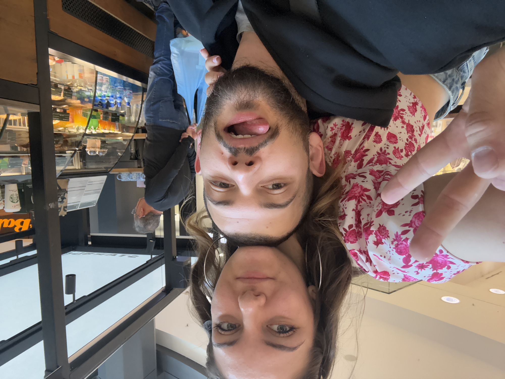 A selfie shows two people, a man and a woman, in what appears to be a public space. The man is above, sticking his tongue out slightly, with a full beard and dark hair. He's wearing a black jacket. Below him is a woman with long hair and large hoop earrings. She has fair skin and appears to be looking directly at the camera. Her clothing is a pink floral print. The background shows a display case containing drinks and pastries, and a glimpse of another person in the background. The exact location within Sofia, Bulgaria, cannot be determined from the image provided.